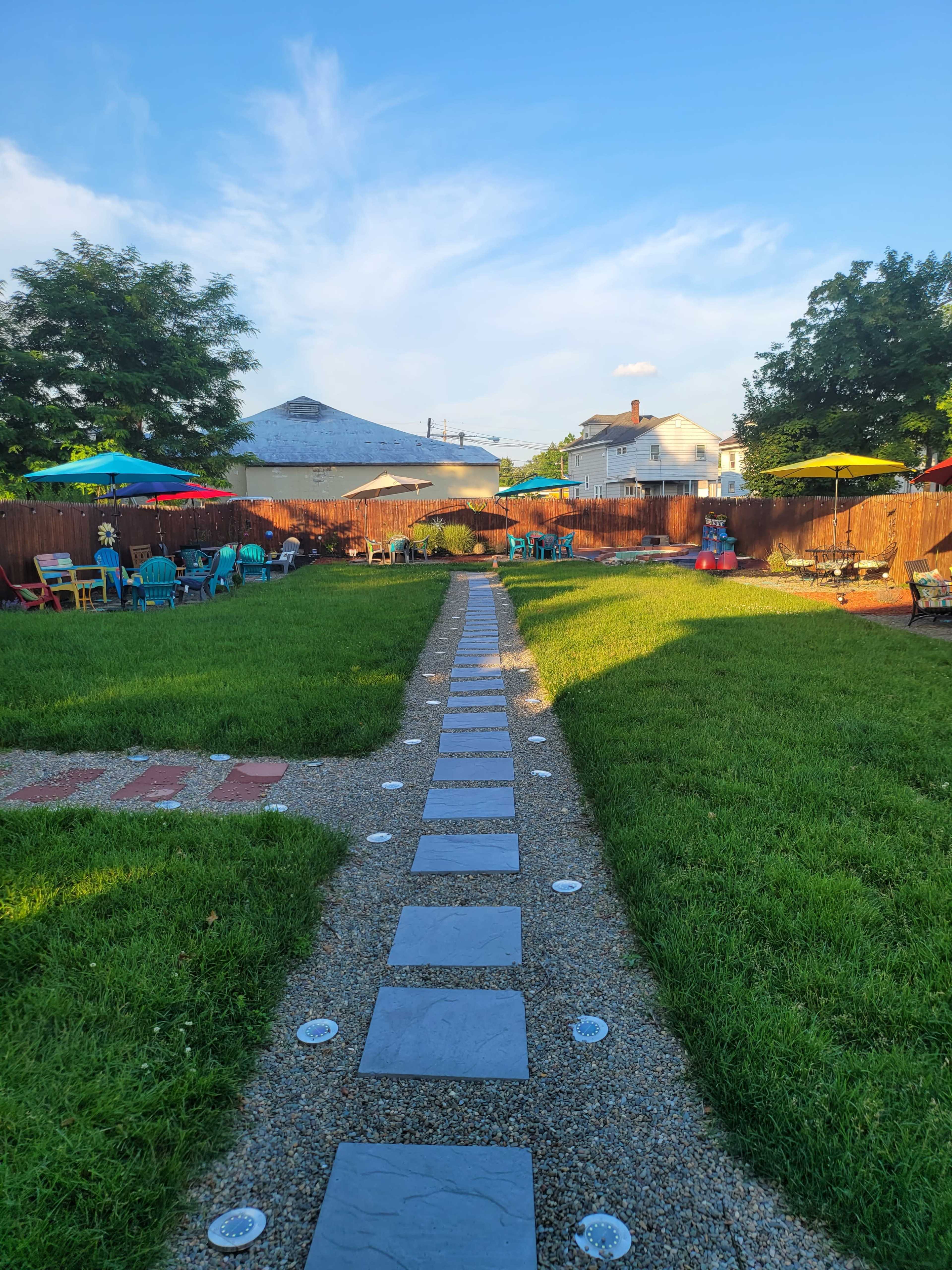A stone pathway lined with grass leads through a backyard featuring colorful umbrellas and seating areas.