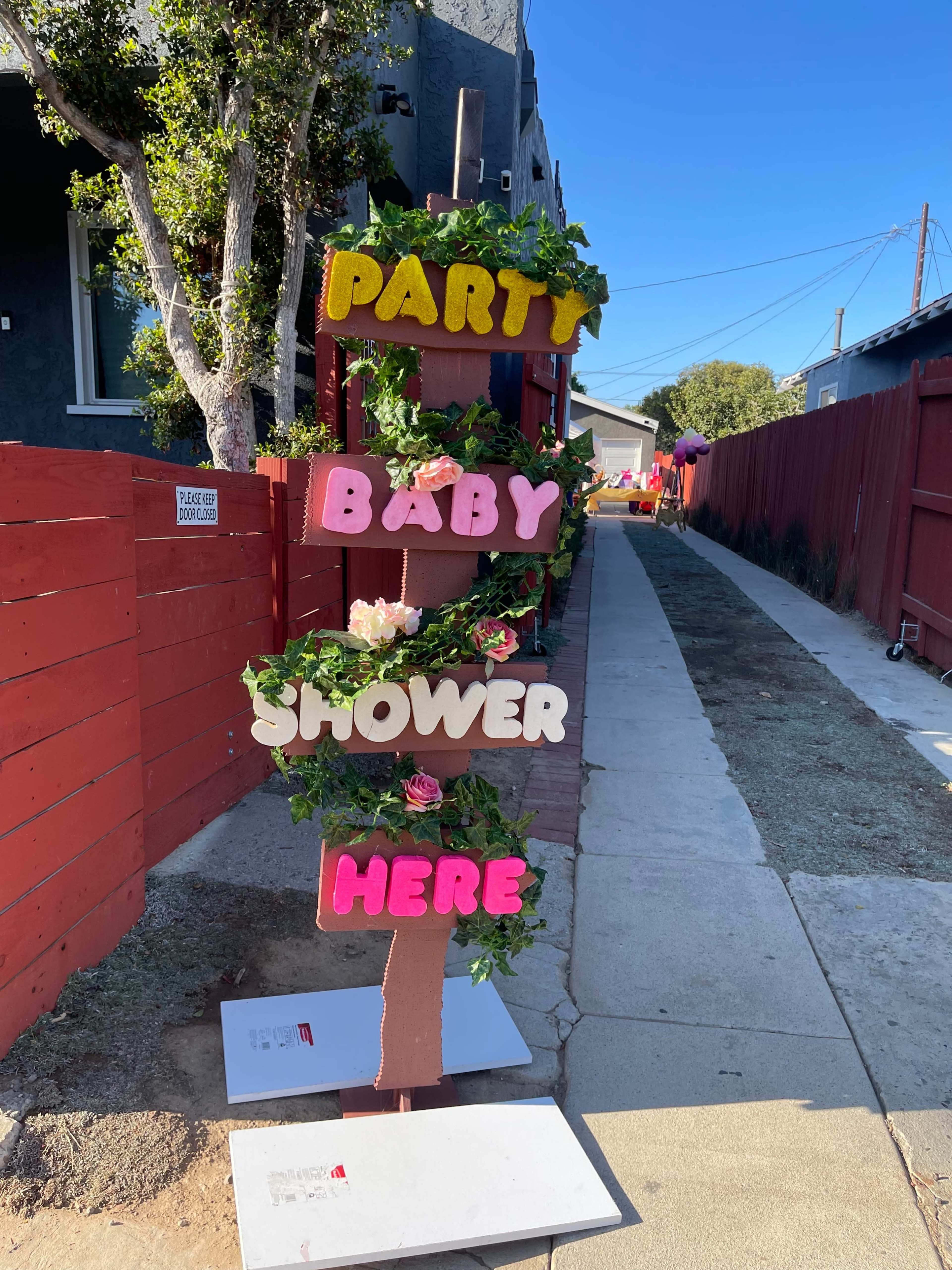 A wooden sign decorated with flowers announces a baby shower, with colorful letters reading "PARTY BABY SHOWER HERE."