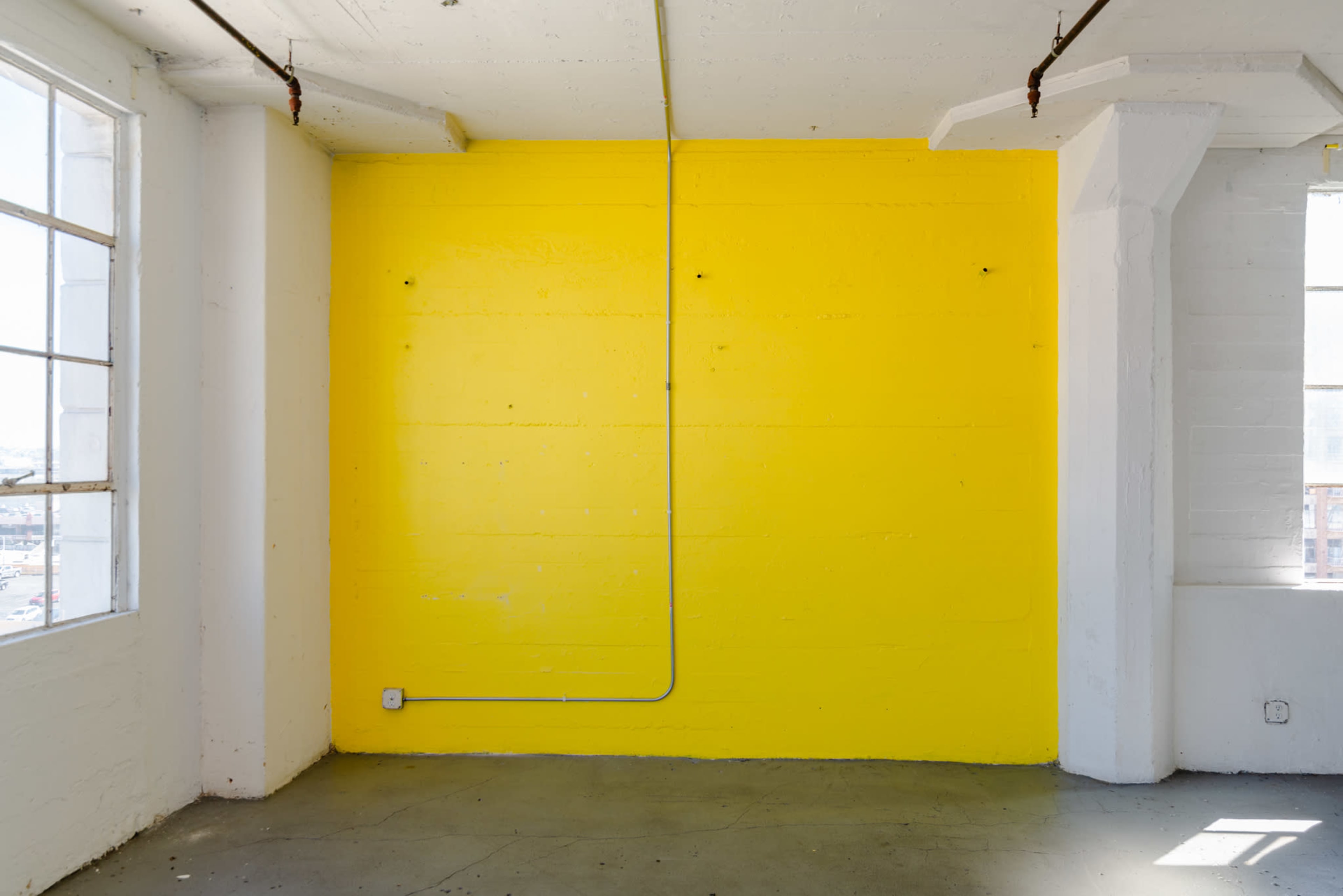 The image shows a bright yellow wall in a spacious room with large windows and exposed pipes.