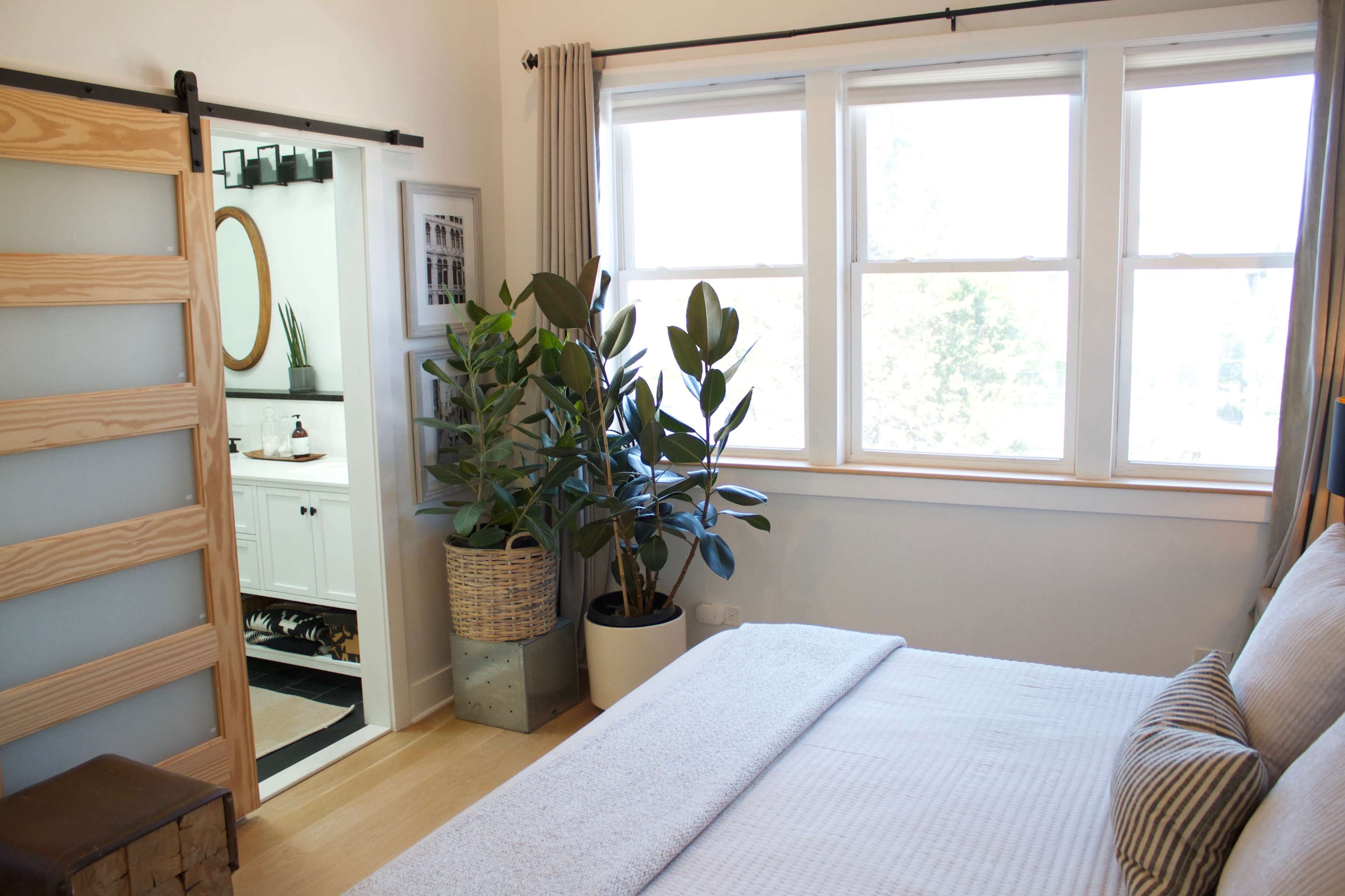 A neatly arranged bedroom features a bed, large windows, and a view of a bathroom with a sliding door and potted plants.