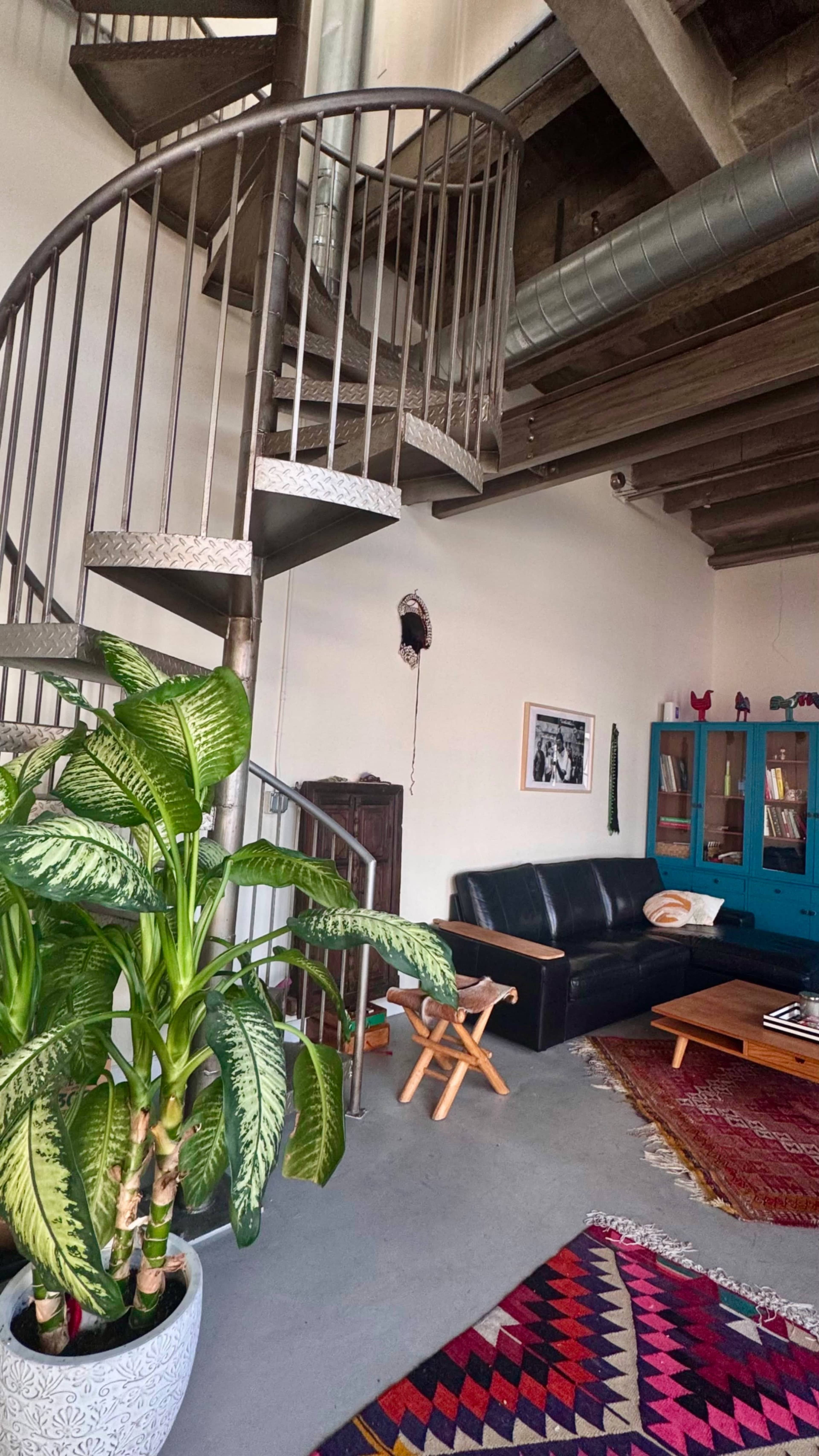 The image shows a spacious living area featuring a spiral staircase, a black leather sofa, a wooden coffee table, and decorative plants.