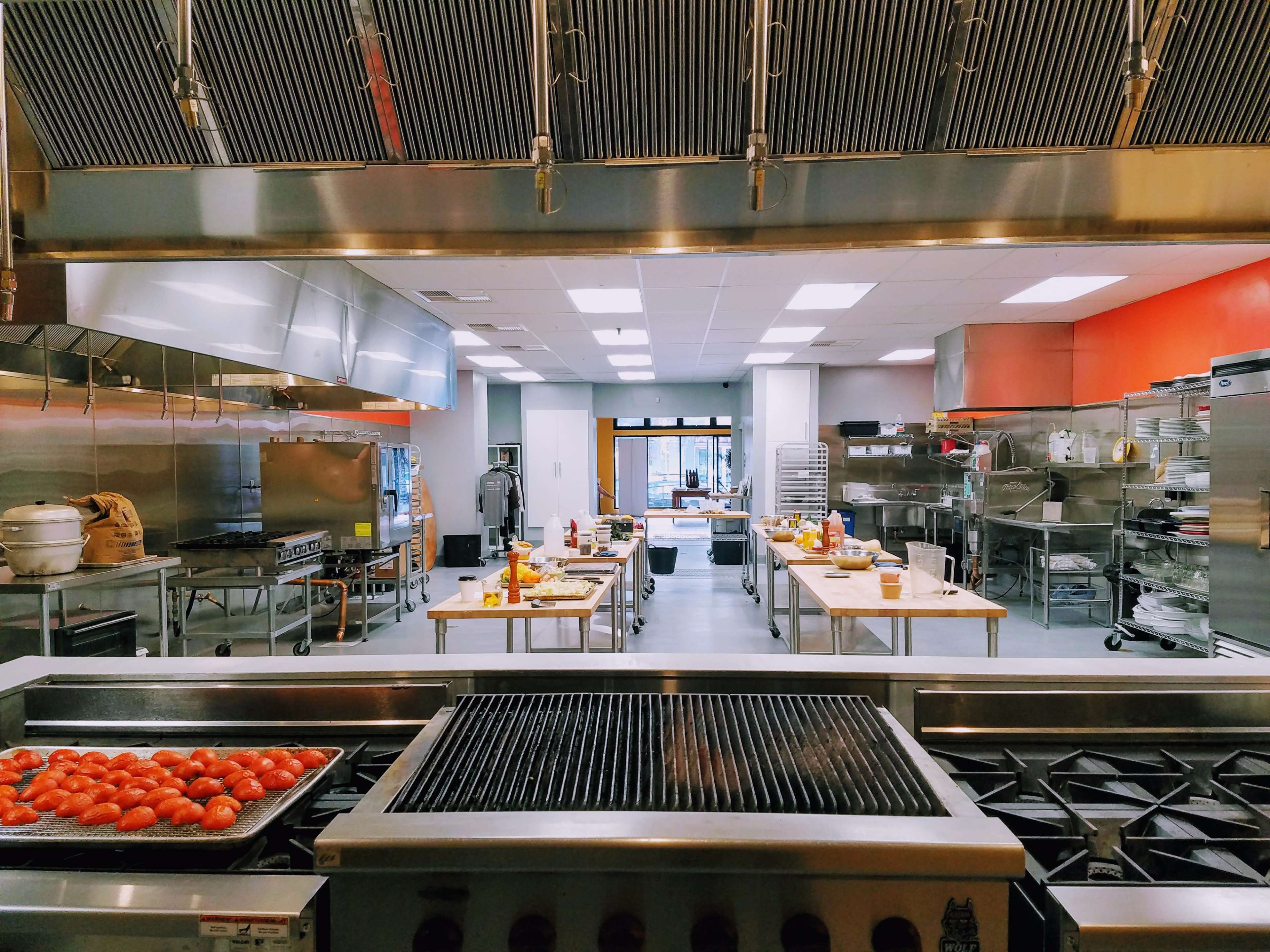 A commercial kitchen is equipped with stainless steel appliances, multiple cooking stations, and large worktables arranged for food preparation.