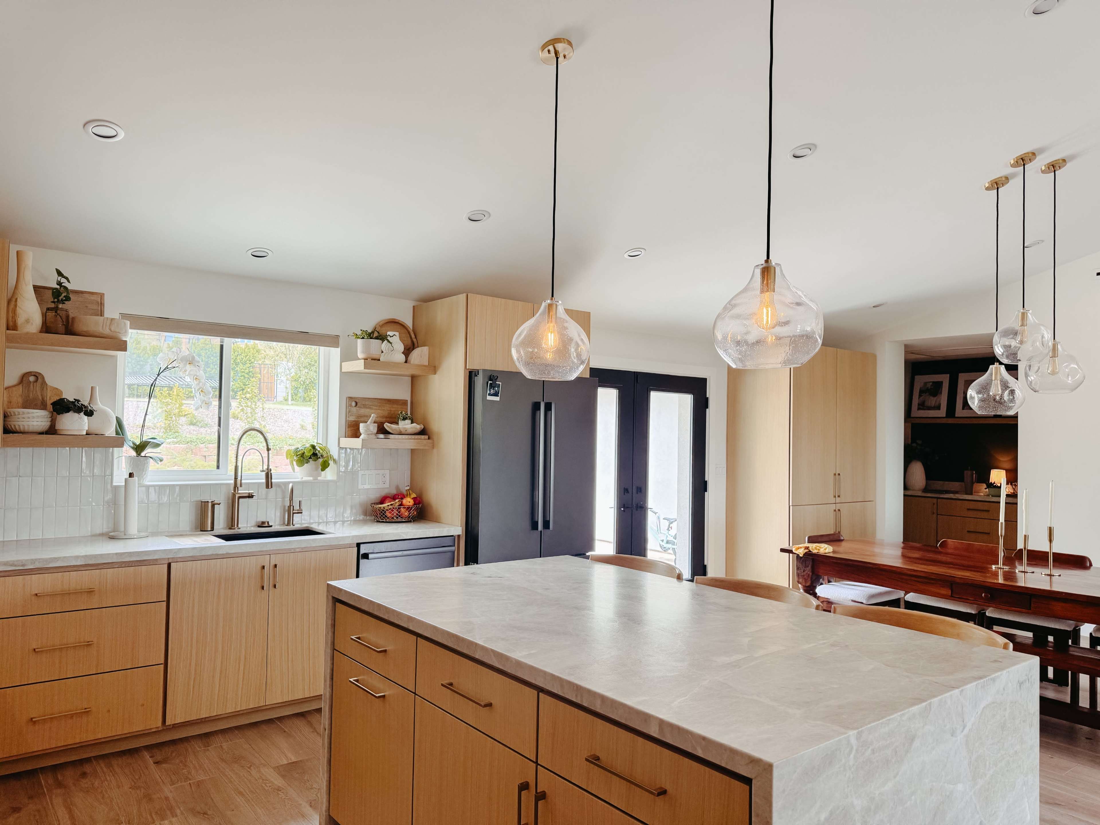 The image shows a modern kitchen with wooden cabinetry, a large island with a stone countertop, and pendant lighting above.
