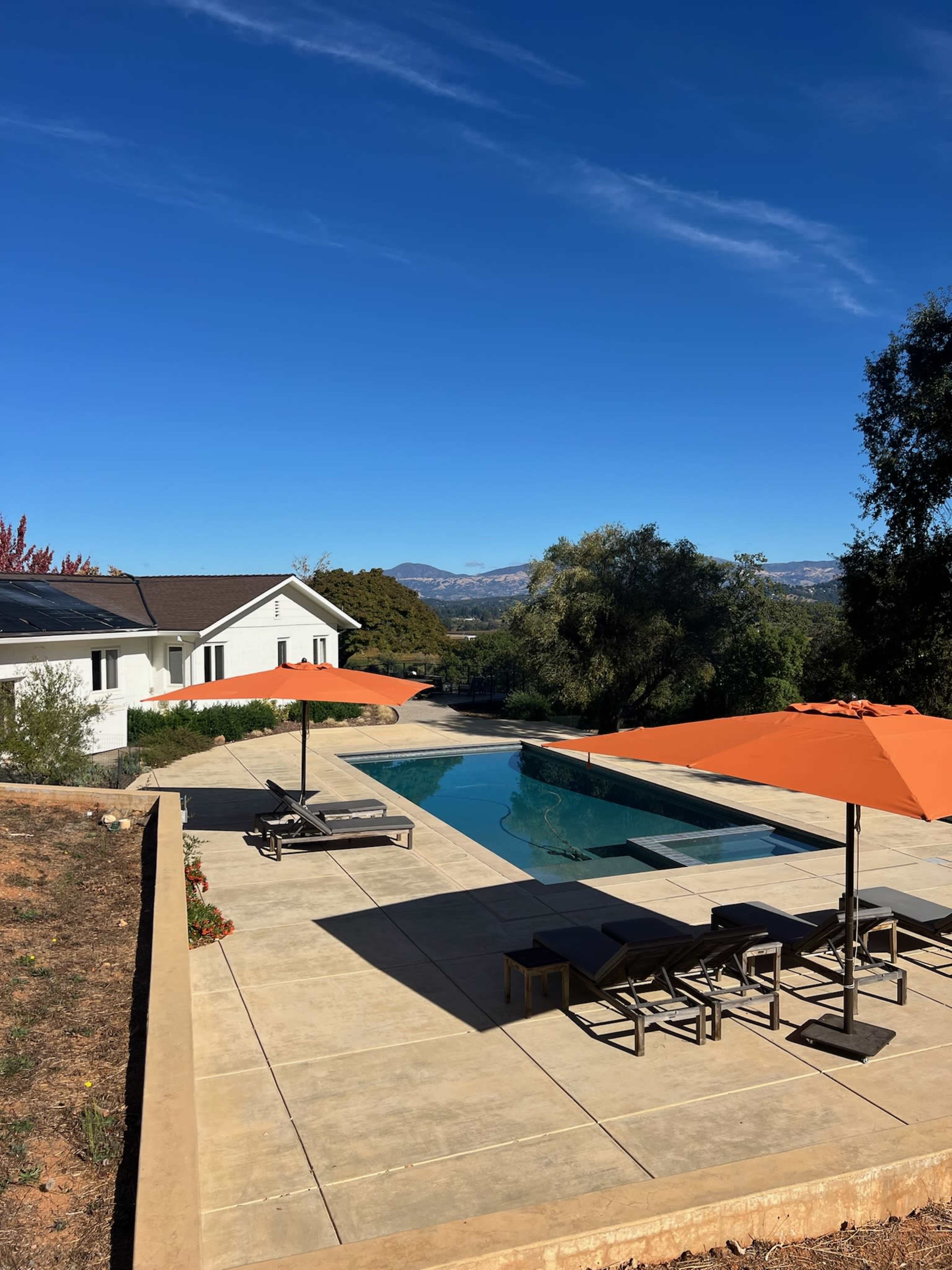 The image shows a modern pool area with orange umbrellas and lounge chairs, set against a backdrop of rolling hills and a clear blue sky.