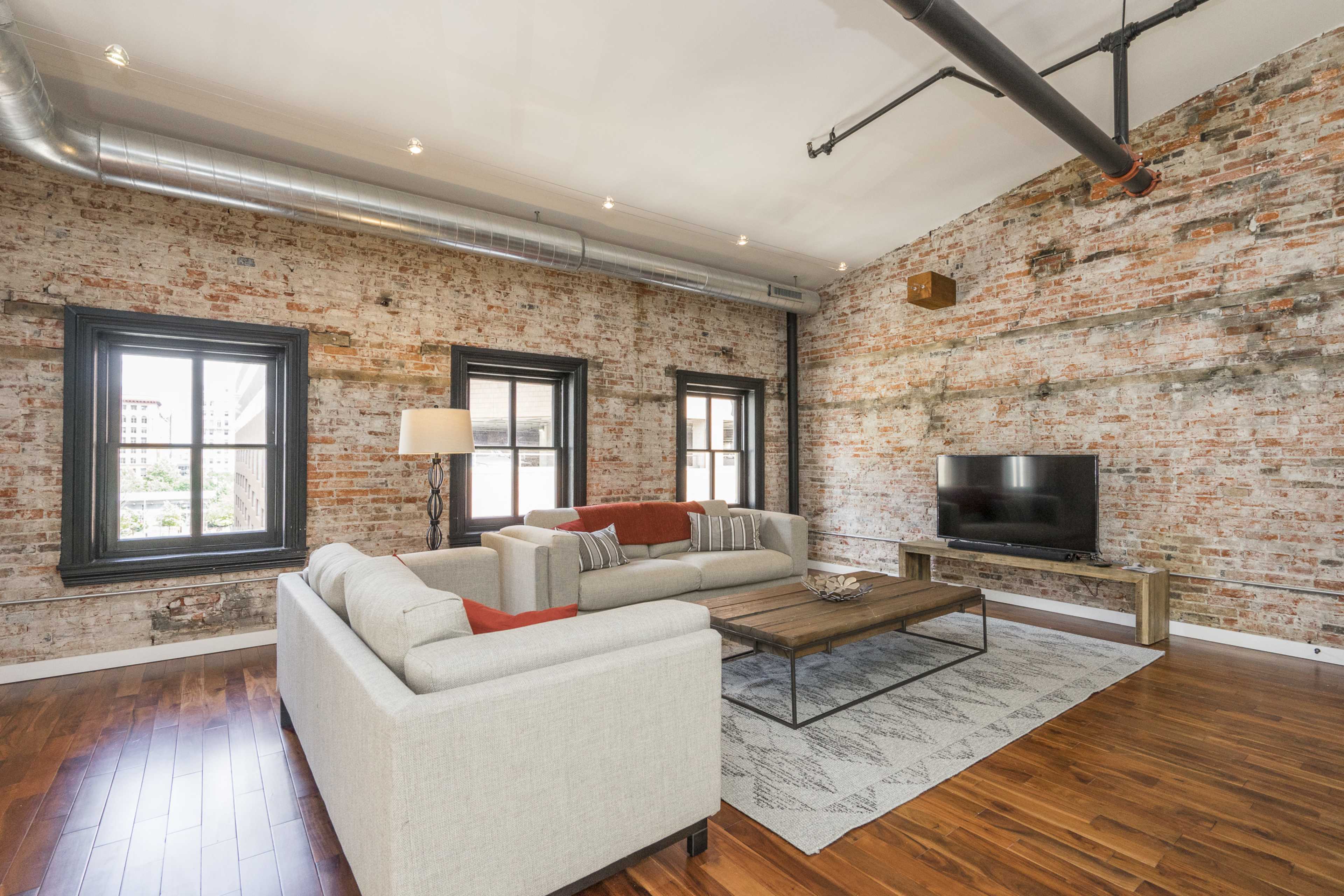 A modern living room with exposed brick walls, hardwood floors, a sectional sofa, a coffee table, and a television mounted on the wall.