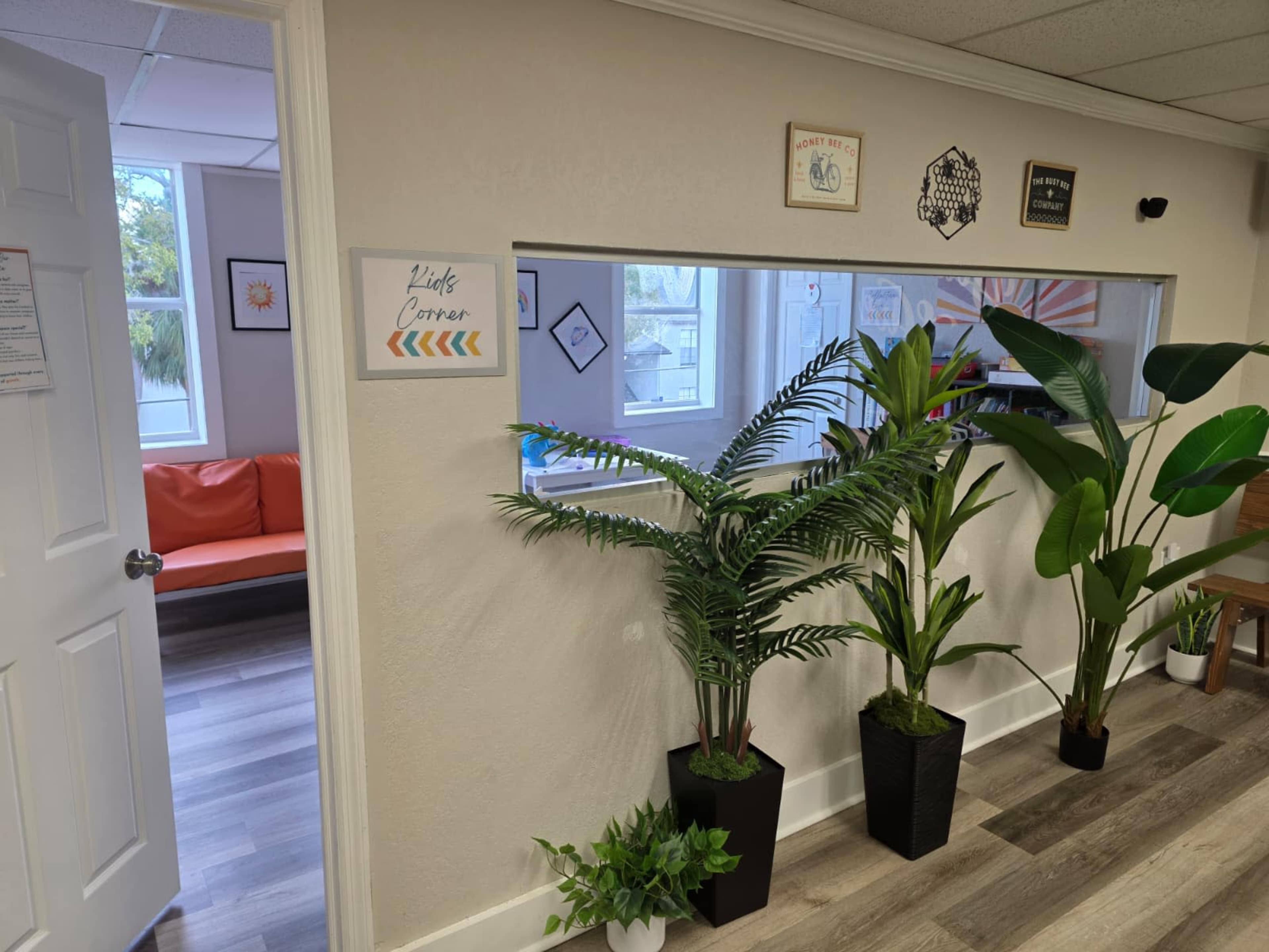 The image shows a room with a large window cutout lined with indoor plants and a sign that reads "Kids' Corner," while in the background, a bright orange couch is visible.