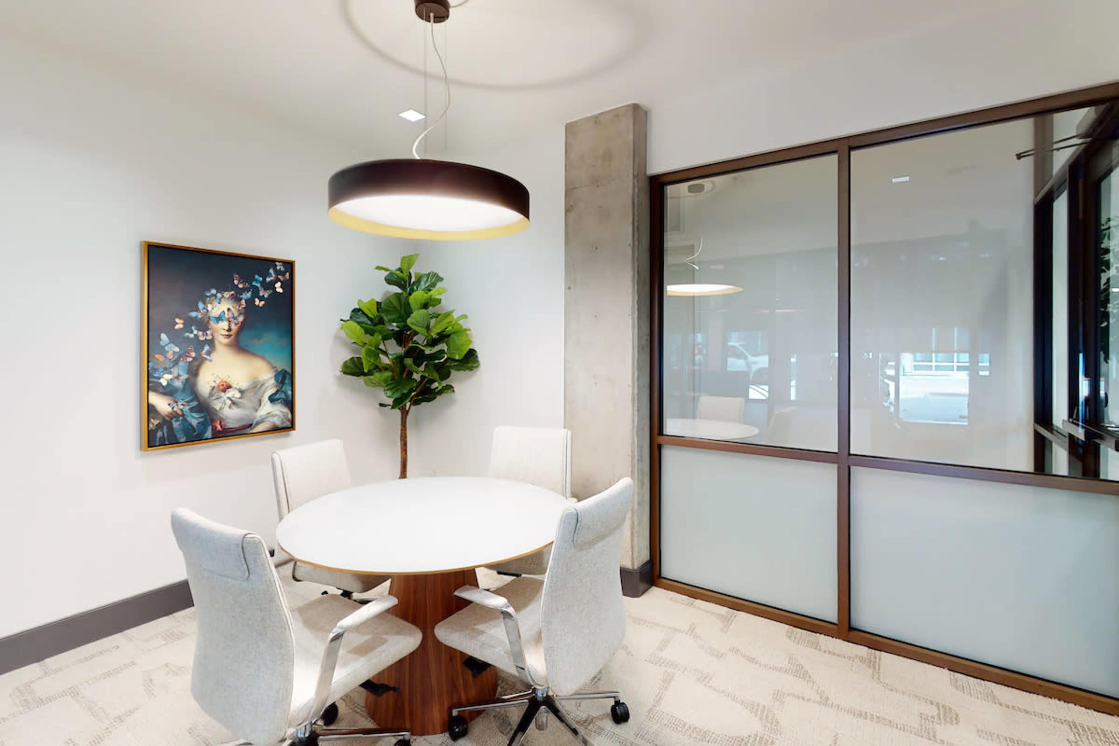 A modern meeting room features a round table surrounded by four chairs, a plant in the corner, and a large artwork on the wall.