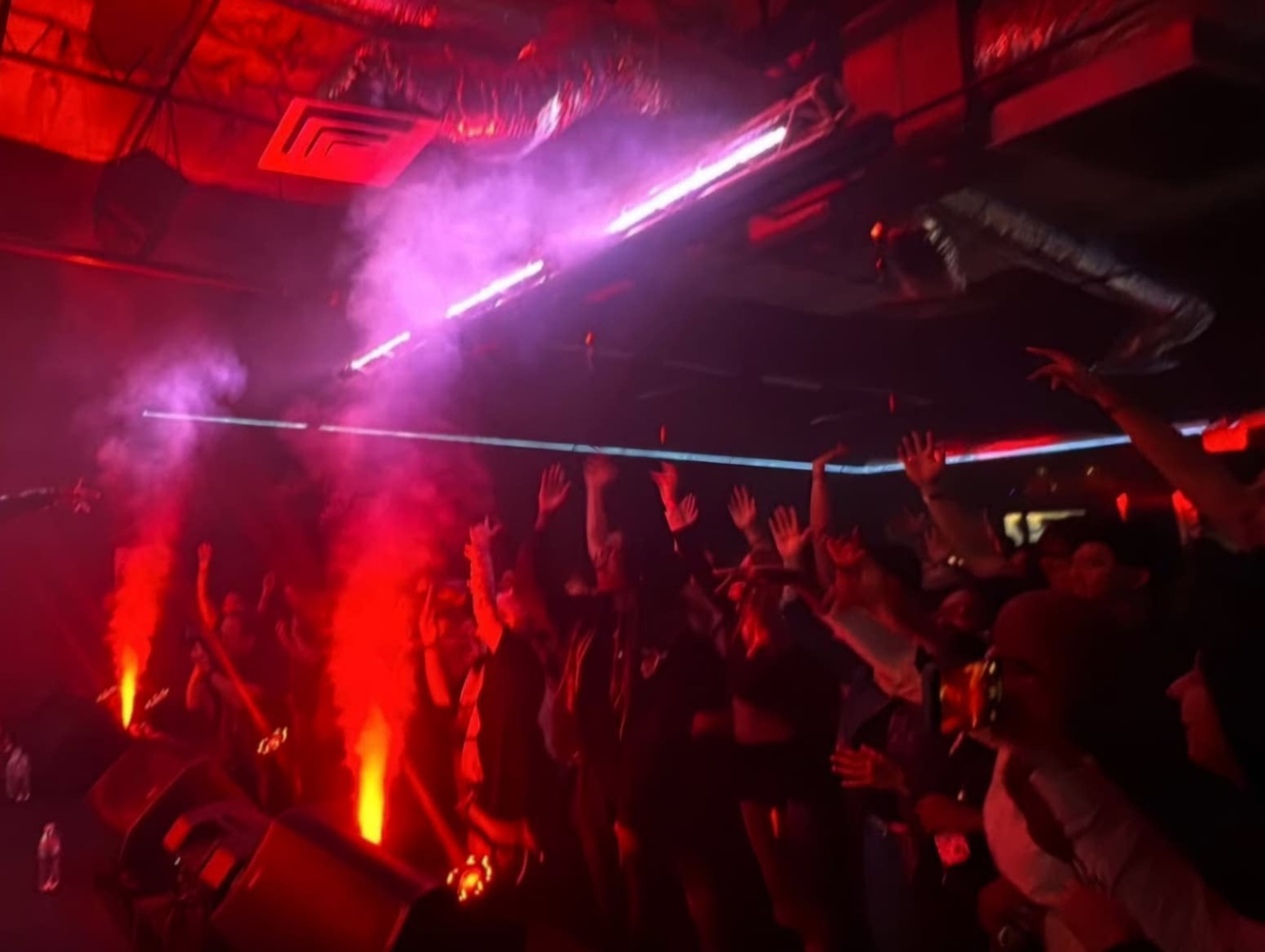 A crowd with raised hands enjoys a performance under red and purple stage lights, with fog and smoke effects in the air.