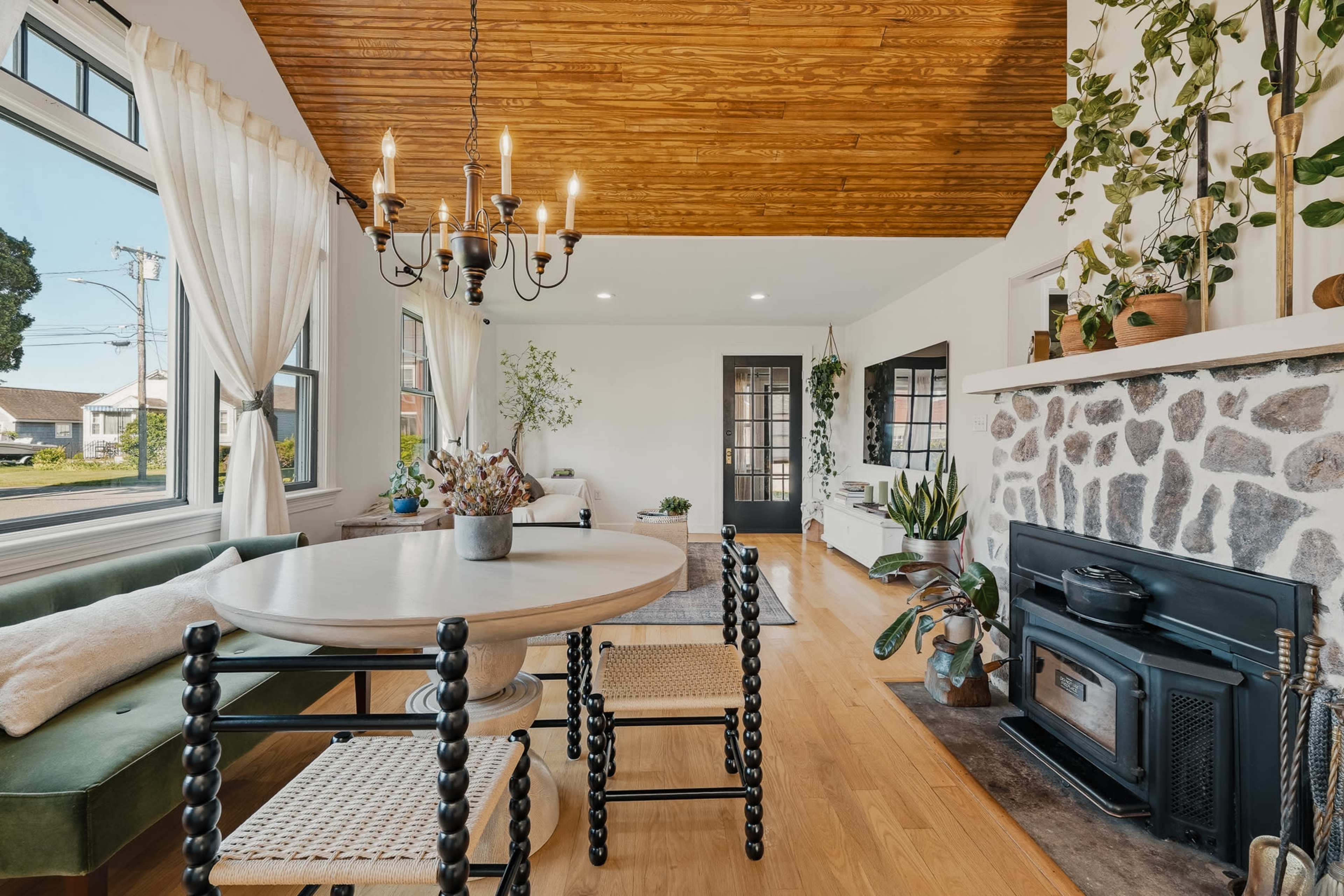 A dining area features a round table surrounded by woven chairs, a stone fireplace, and a large window that lets in natural light.
