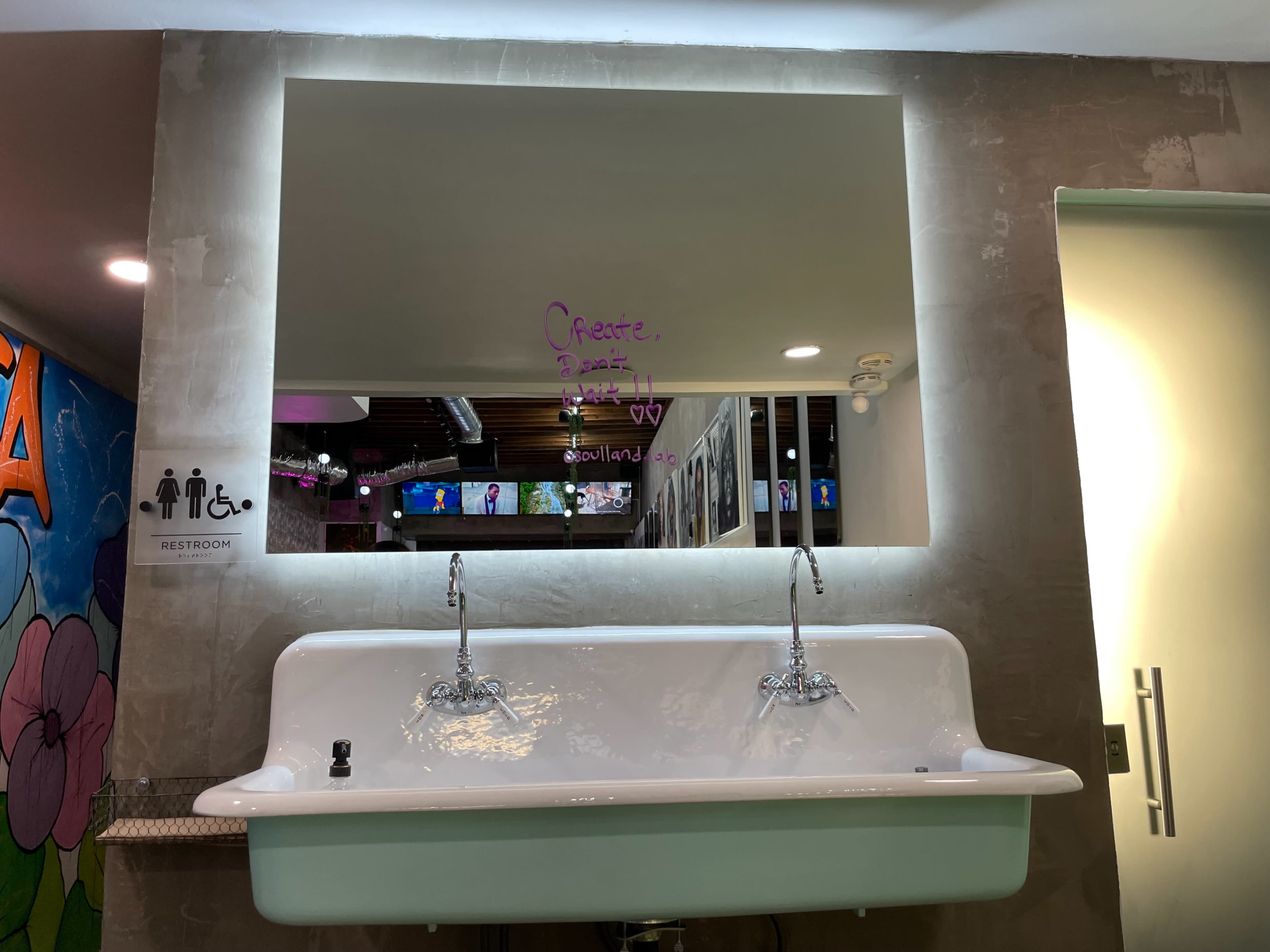 A restroom features a large sink beneath a illuminated mirror with hand-written text and a modern aesthetic.