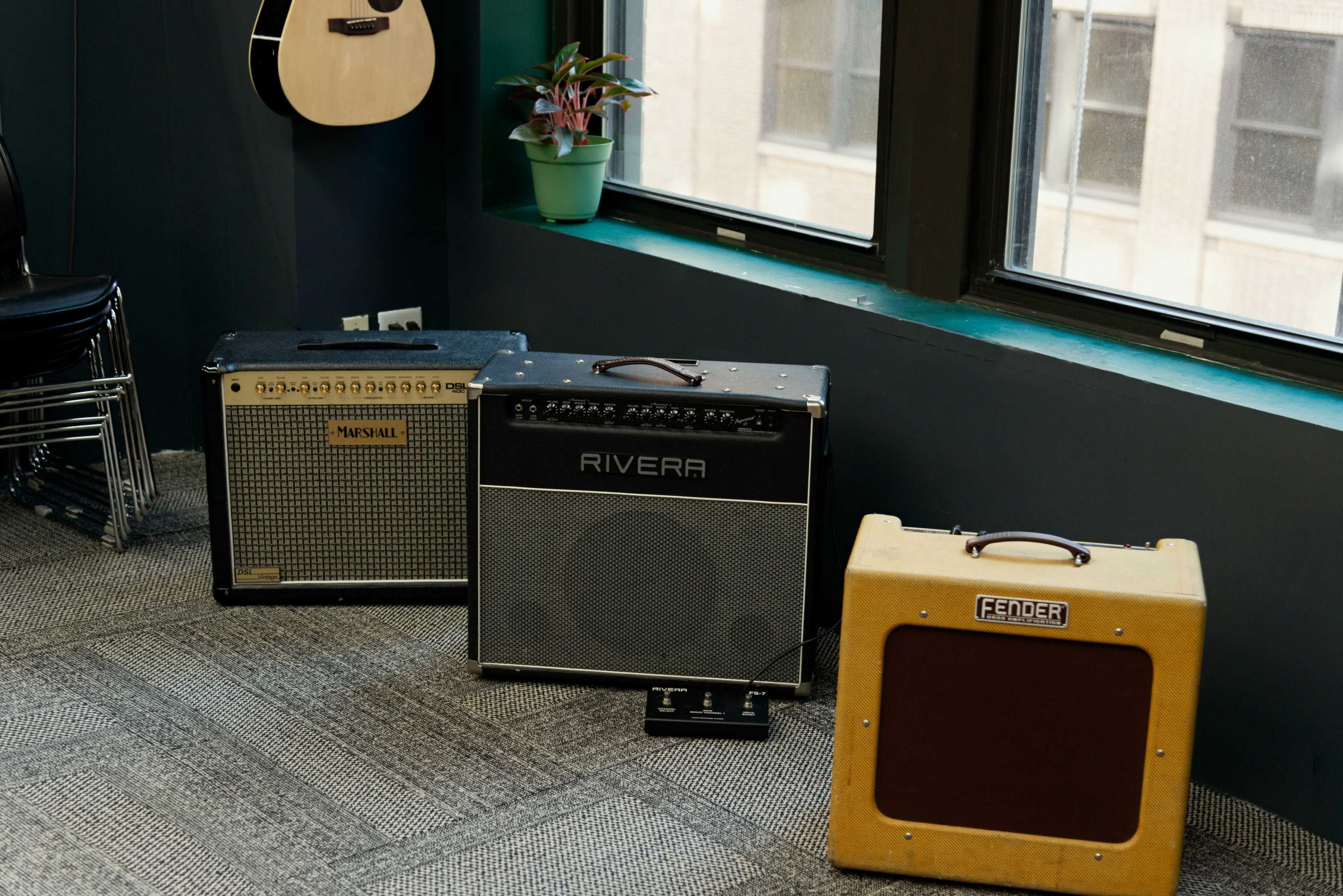 The image shows three guitar amplifiers placed on a carpeted floor near a window, with a potted plant and a guitar hanging on the wall.
