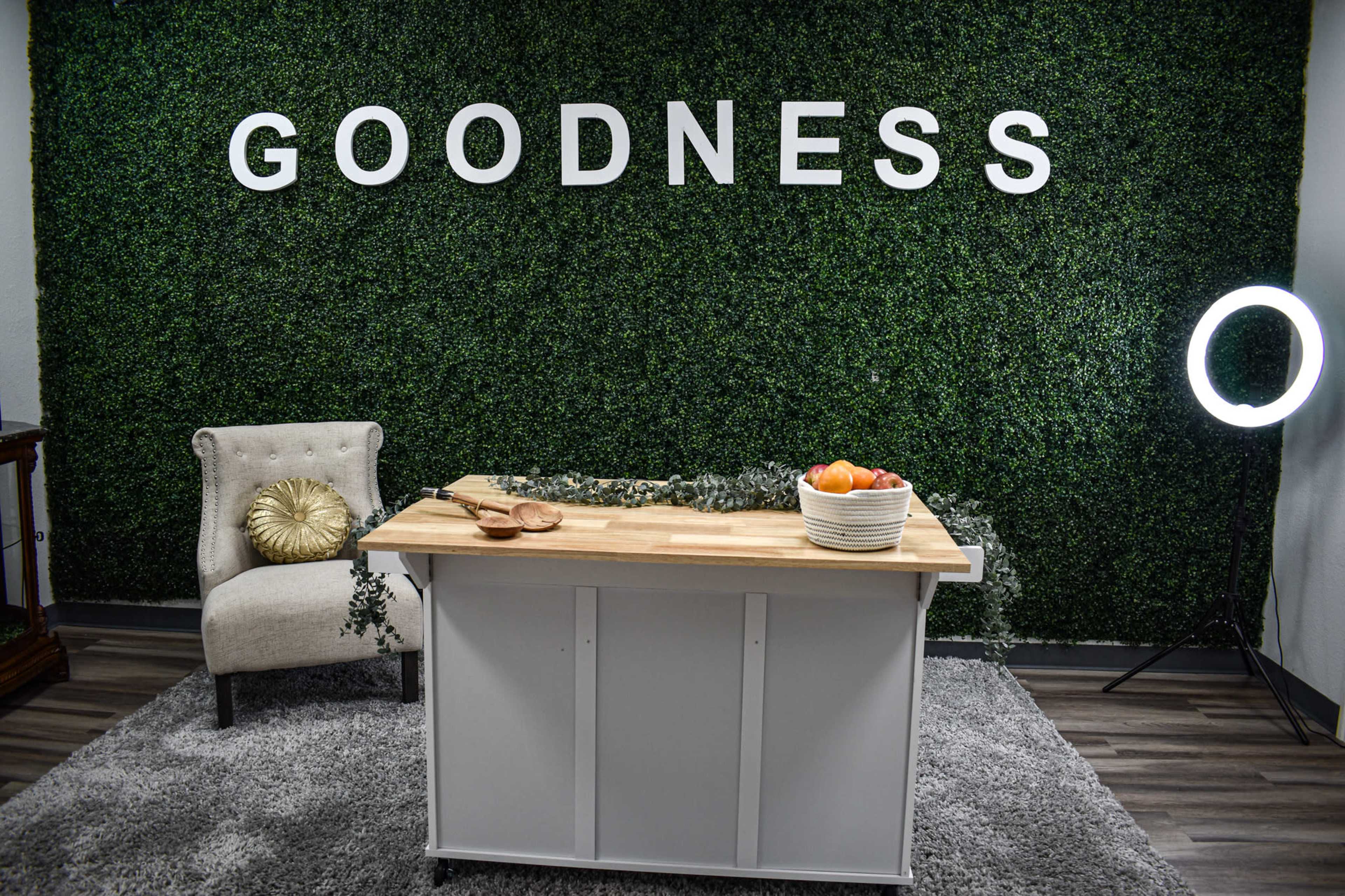 A wooden table with a bowl of fruit is positioned in front of a green wall adorned with the word "GOODNESS" in large letters, with a chair and a ring light nearby.