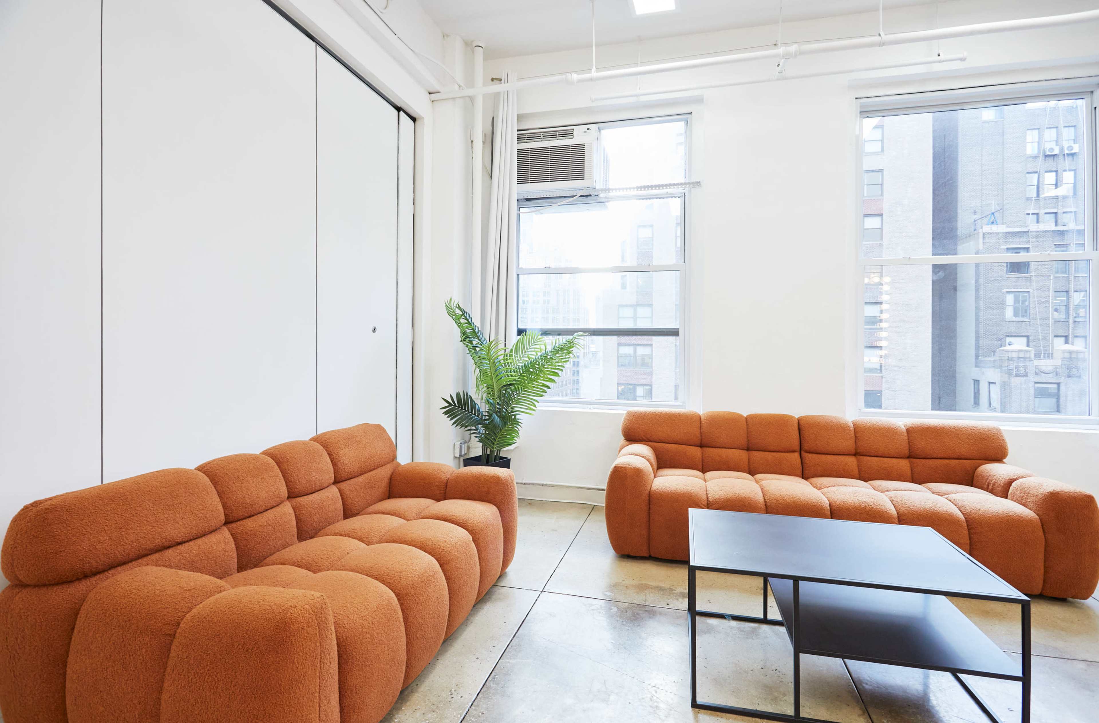 The image shows a modern office space with two orange sofas, a black coffee table, and large windows providing natural light.