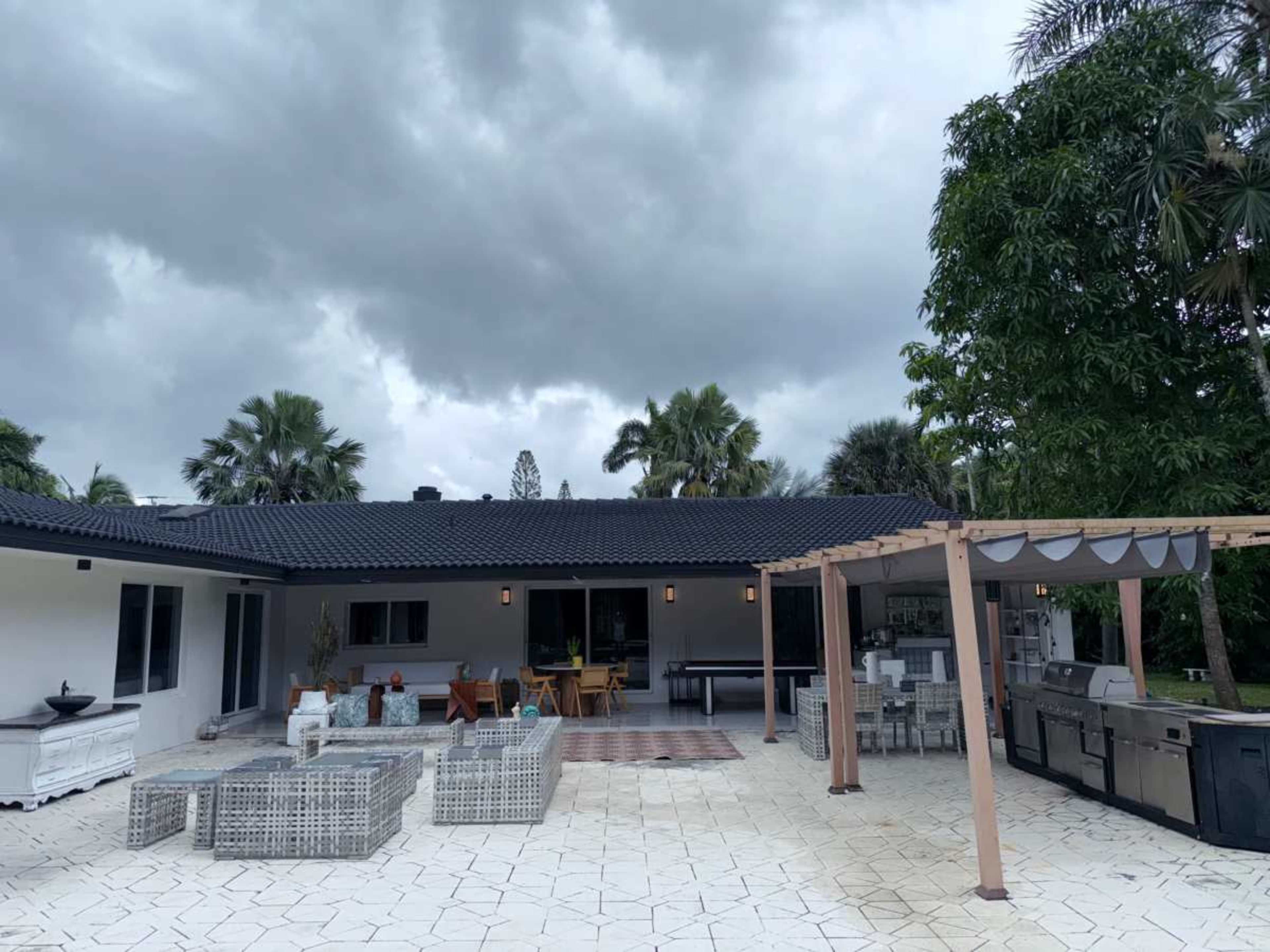 A modern one-story house with a patio area featuring outdoor seating and a built-in grill, surrounded by palm trees under an overcast sky.