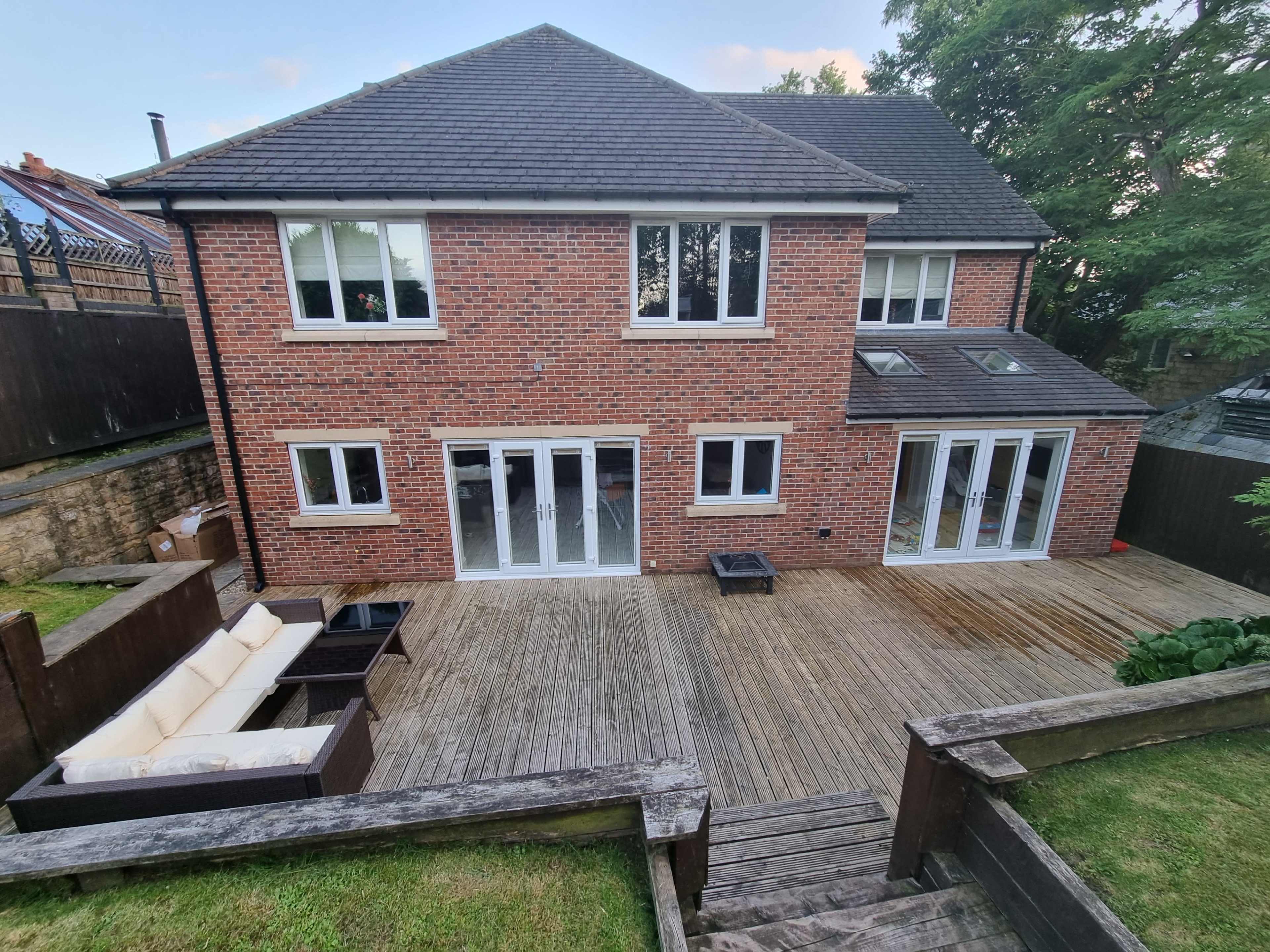 A two-story brick house with multiple windows, a wooden deck, and outdoor seating, surrounded by greenery.