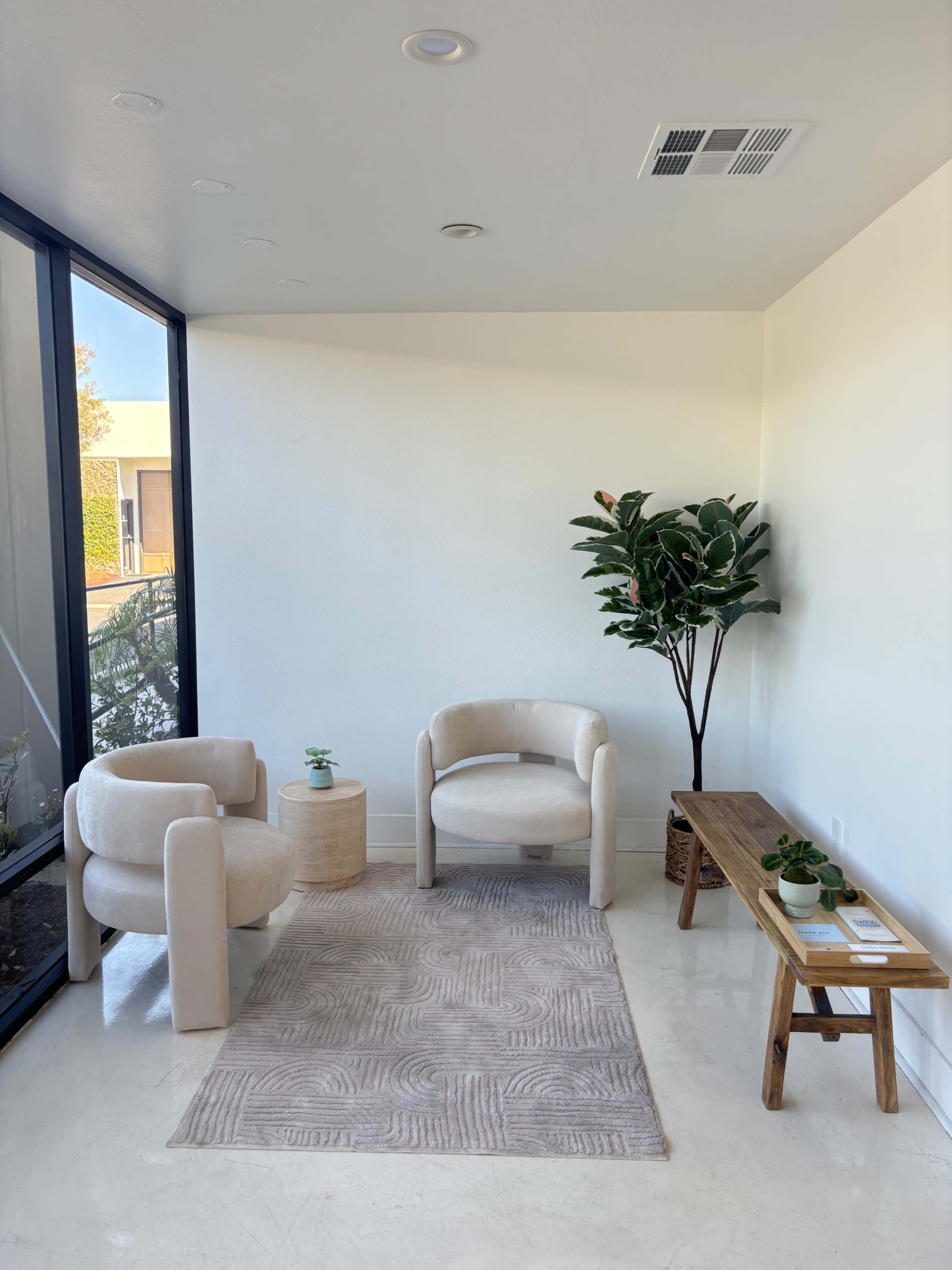 The image shows a minimalist corner of a room with two beige armchairs, a small round table, a wooden bench, and a potted plant, all arranged on a light rug.