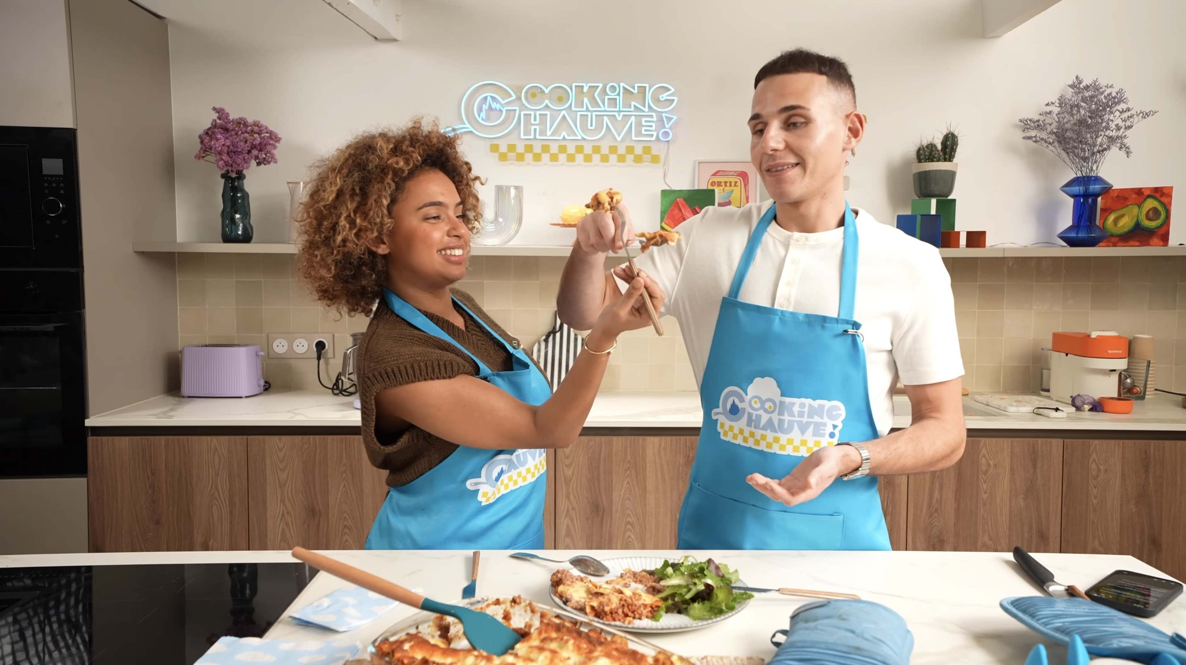 Two individuals in matching blue aprons are playfully holding up food items in a modern kitchen with various cooking ingredients and utensils visible.