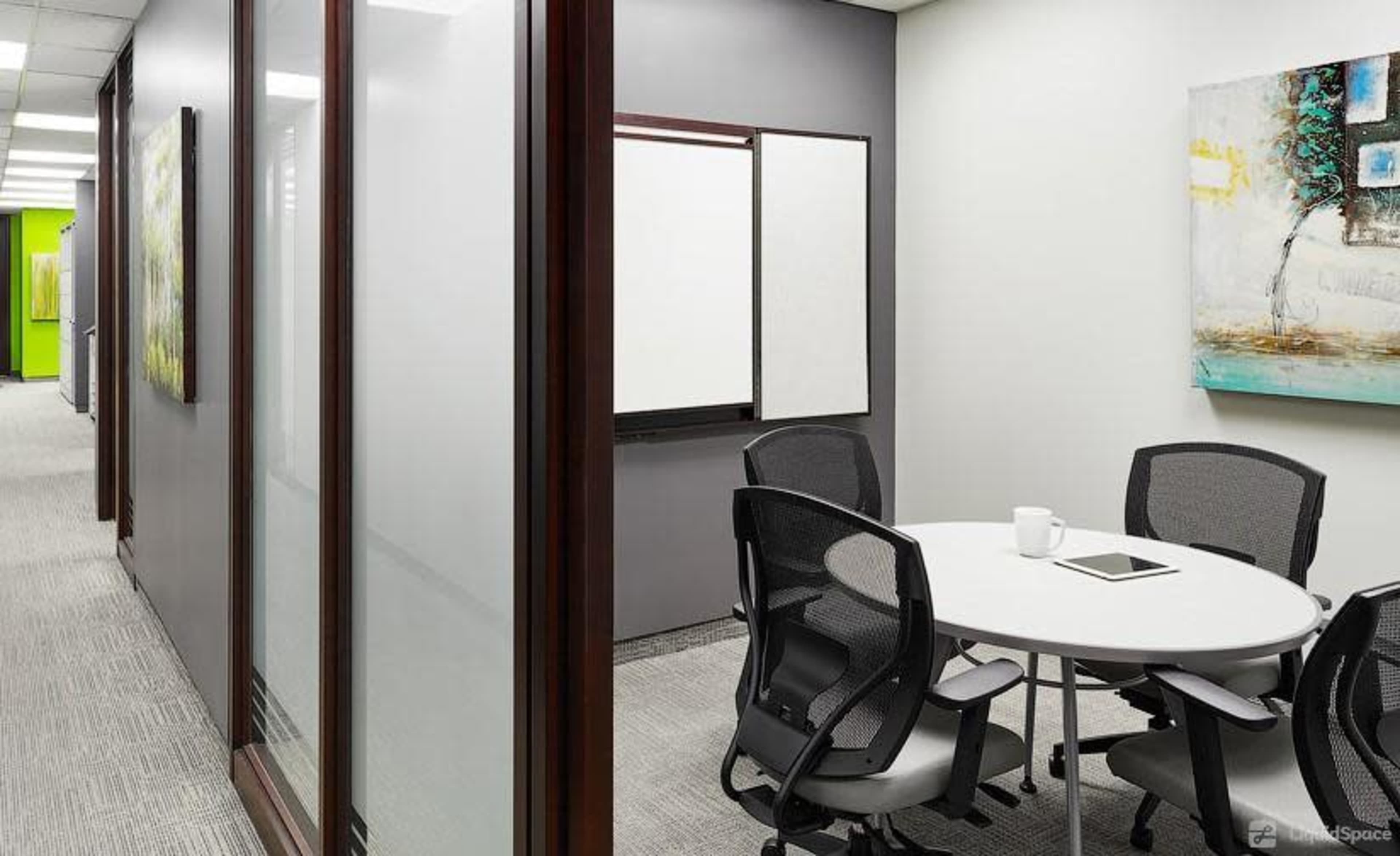 A modern office meeting room with a round table and chairs, flanked by glass walls and a whiteboard.