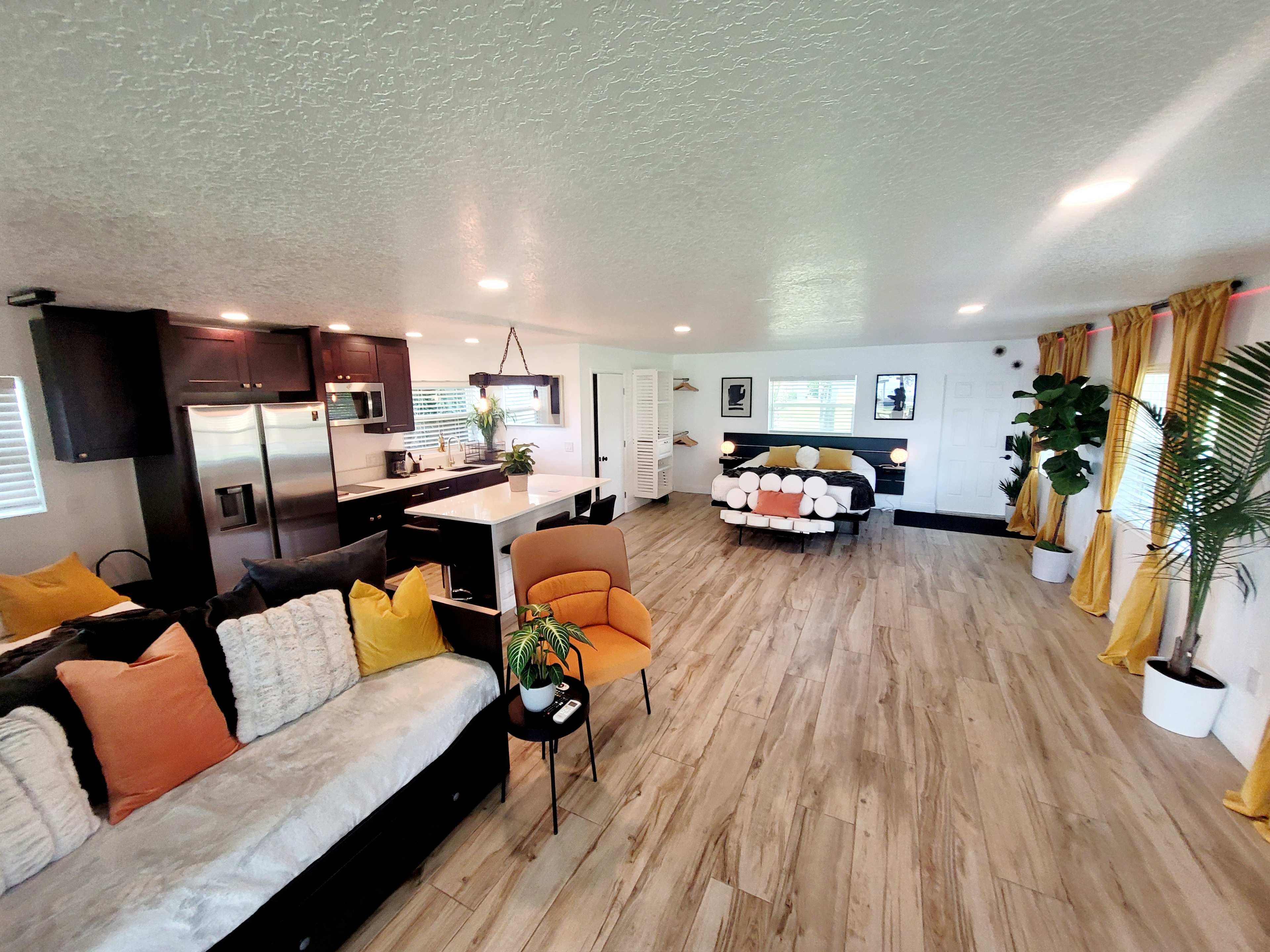 A modern living space with a seating area, a kitchen in the background, and large windows allowing natural light to enter.