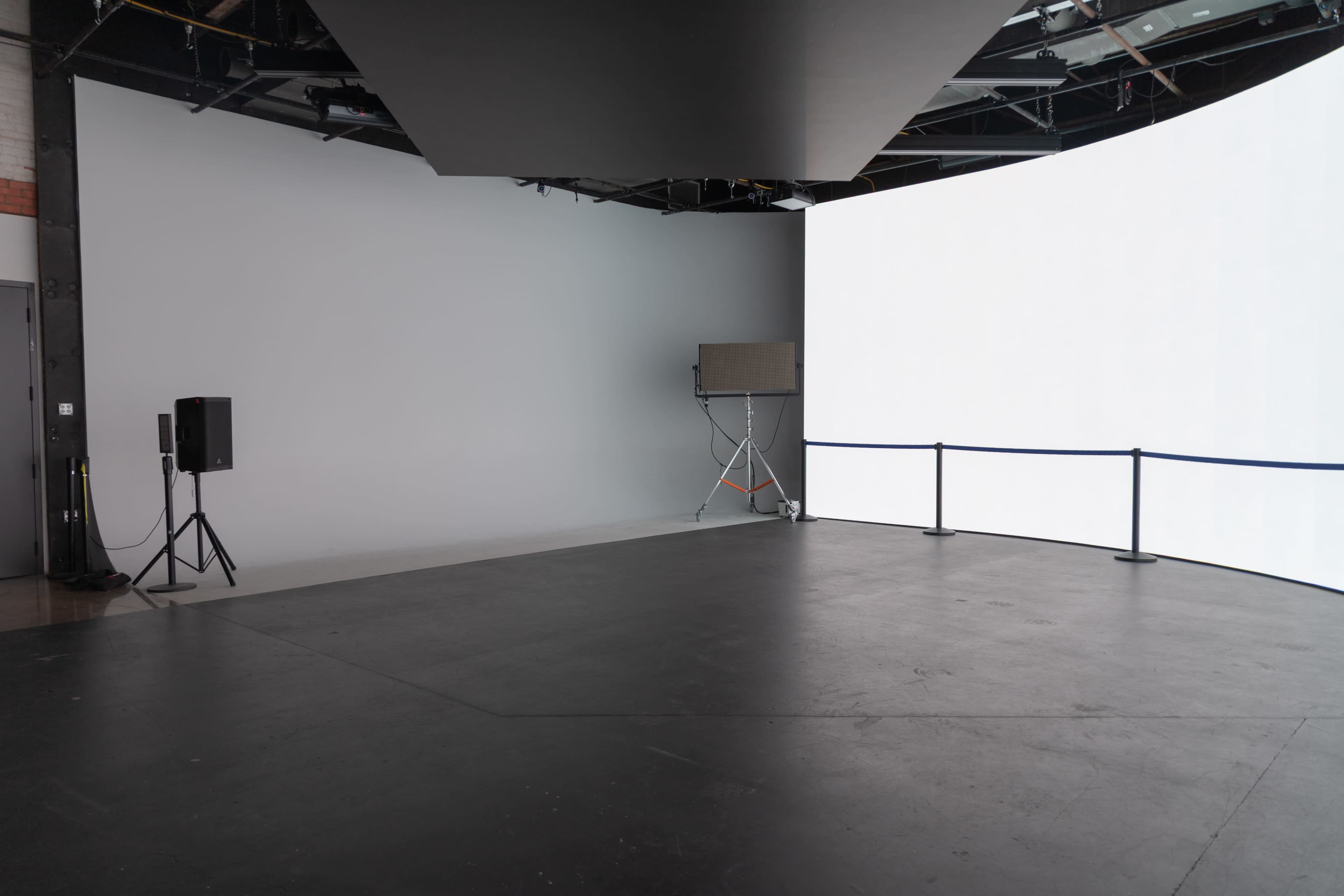 The image shows a studio space with a curved backdrop and a large illuminated white screen on one side, along with a speaker and safety barriers.