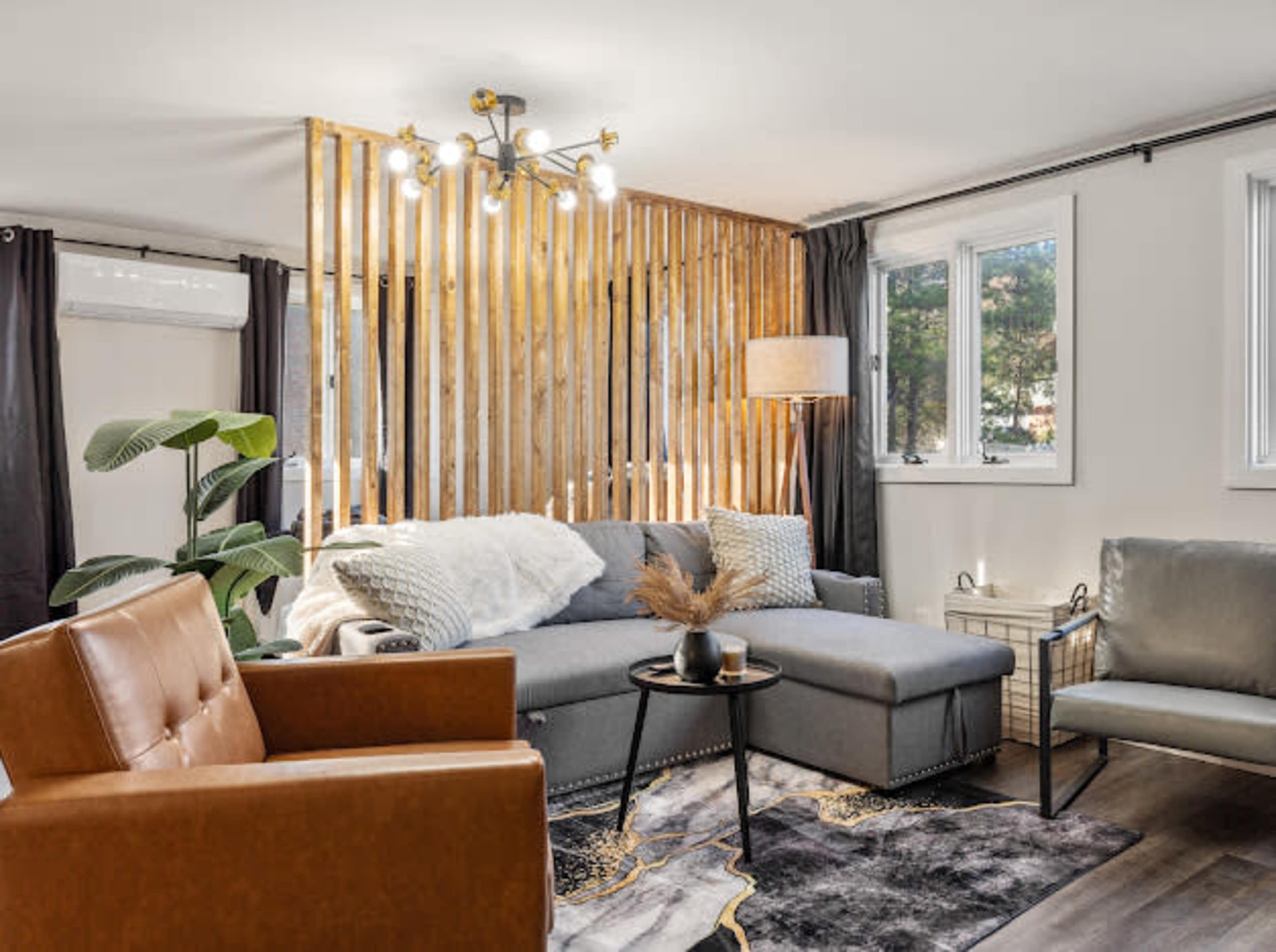 The living room features a sectional sofa, a round coffee table, and a wooden slatted wall, with large windows allowing natural light.