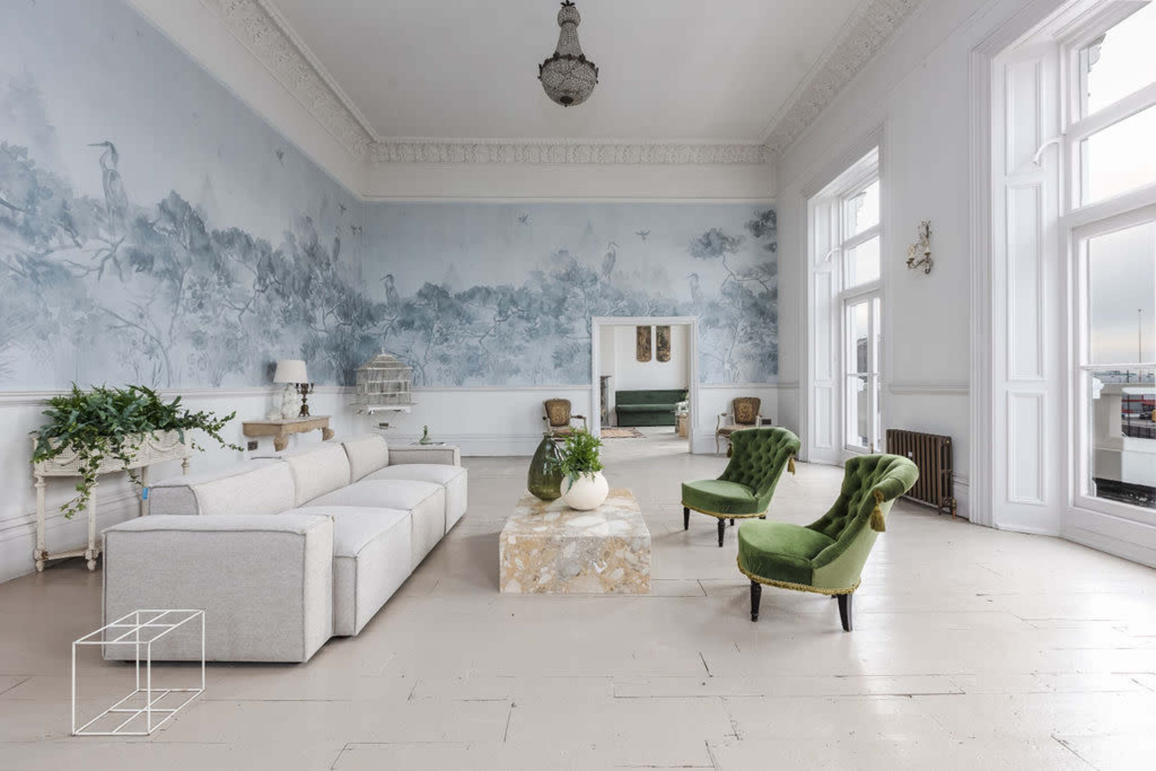 The spacious living room features a light-colored sectional sofa, two green velvet chairs, a marble coffee table, and a large mural depicting a nature scene.