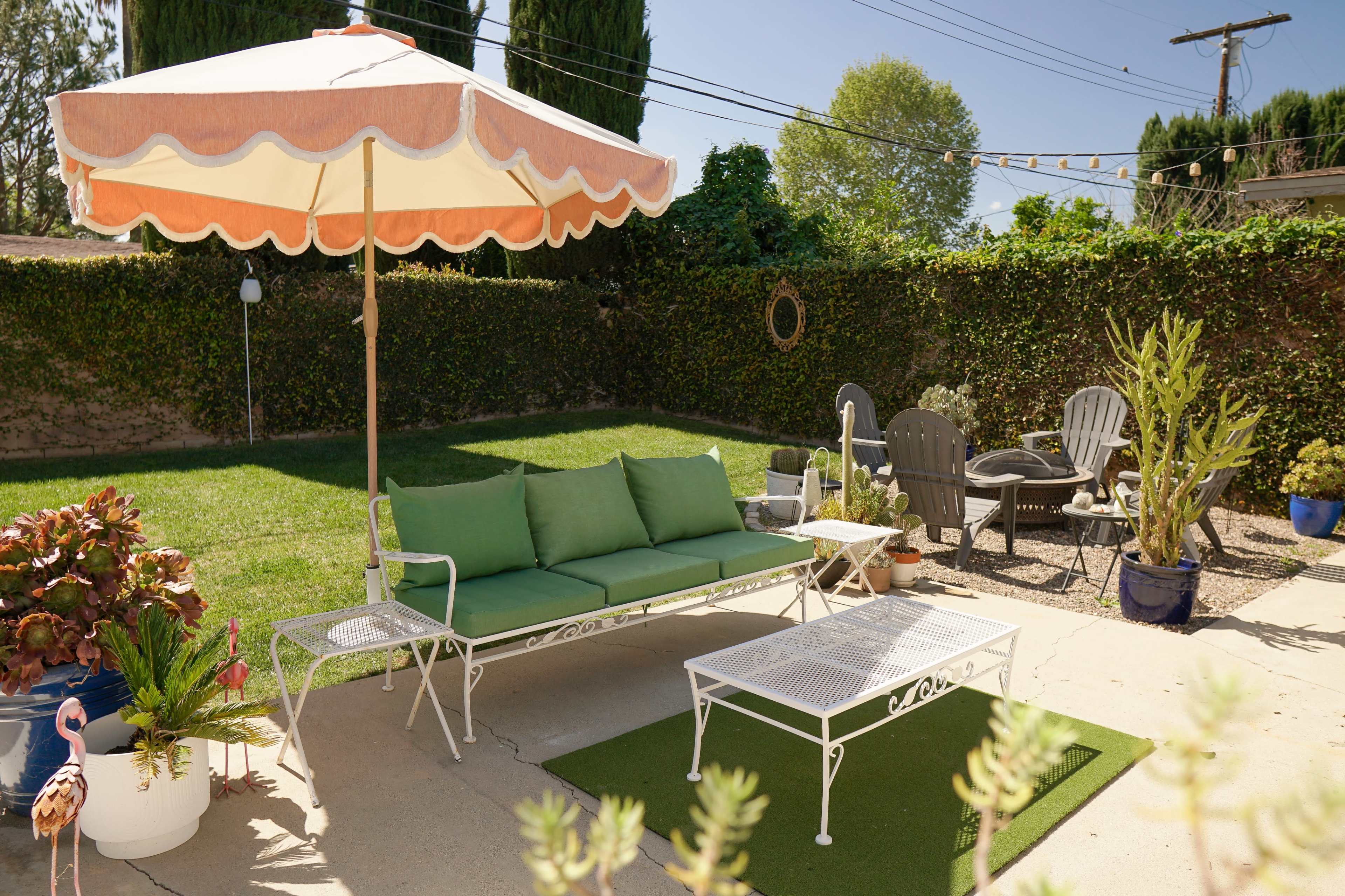 A well-manicured backyard features a green sofa set under a large umbrella, surrounded by potted plants and outdoor seating.