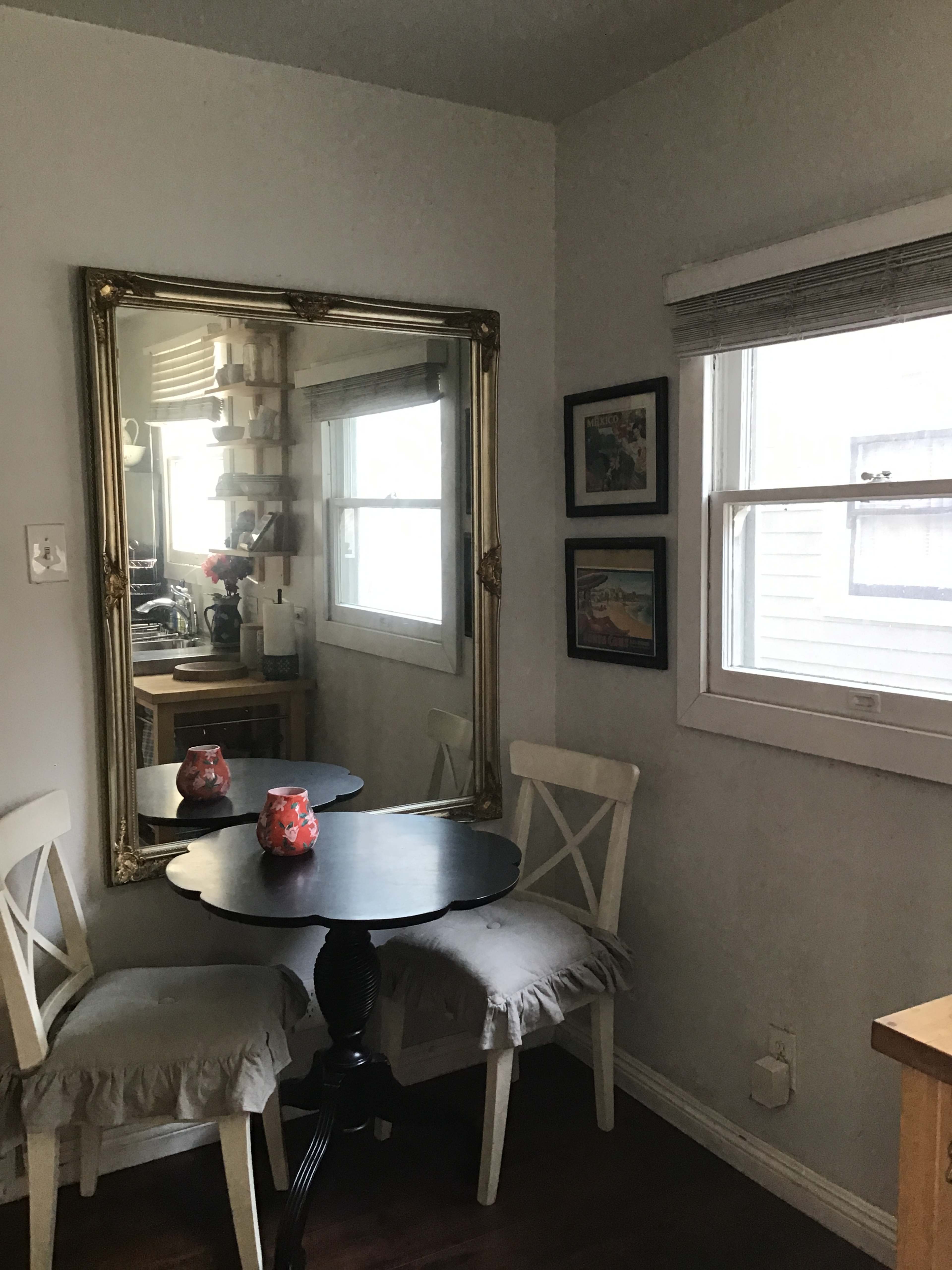 A small dining area with a round black table, two white chairs, a large mirror on the wall, and a window with blinds.
