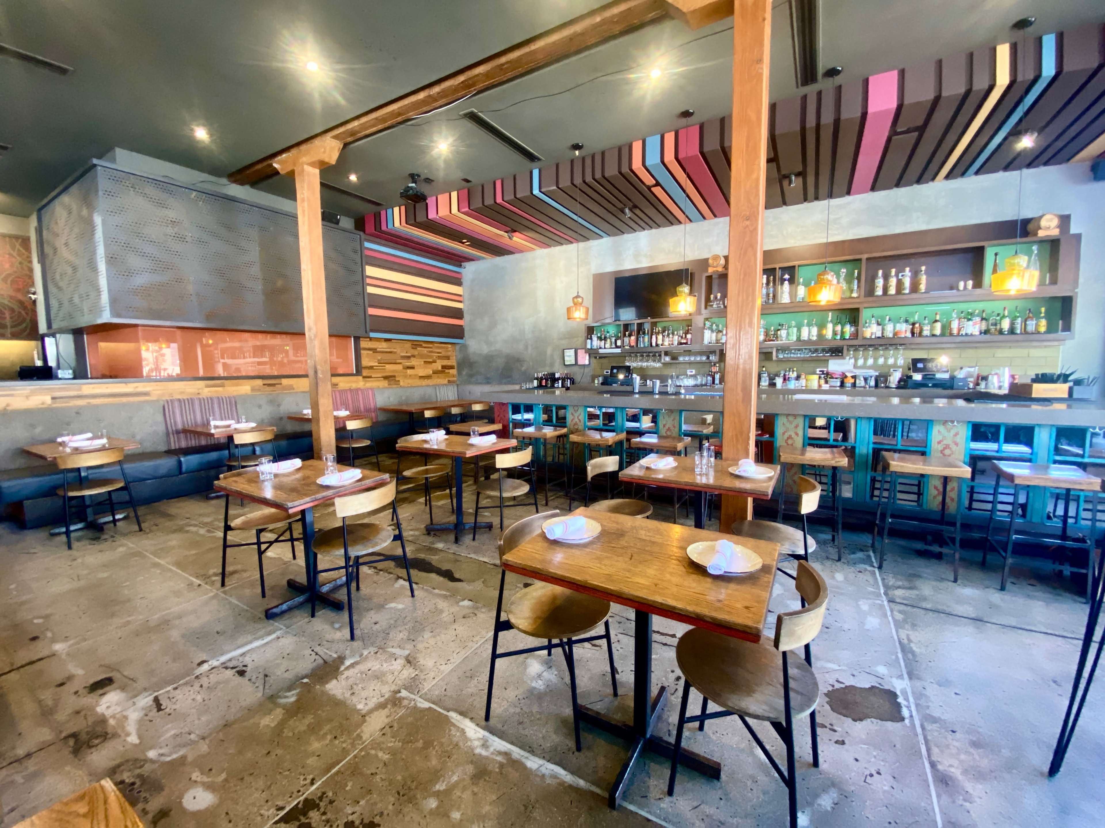 A modern restaurant interior with wooden tables and chairs, a bar area in the back, and colorful striped overhead decorations.