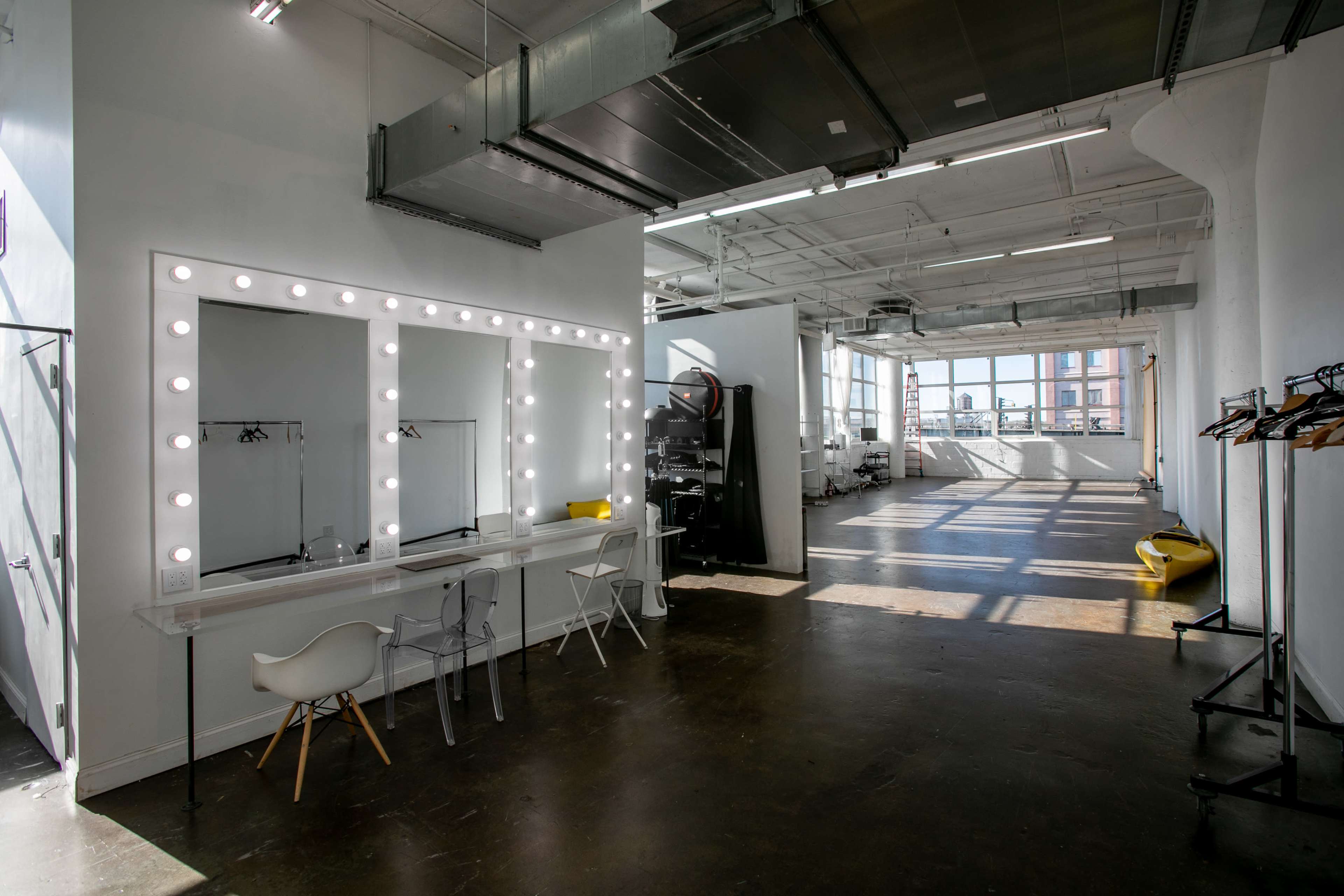 A spacious bright room featuring a large vanity mirror with lights, a white table with chairs, and various furniture pieces against the walls, all illuminated by natural light from wide windows.
