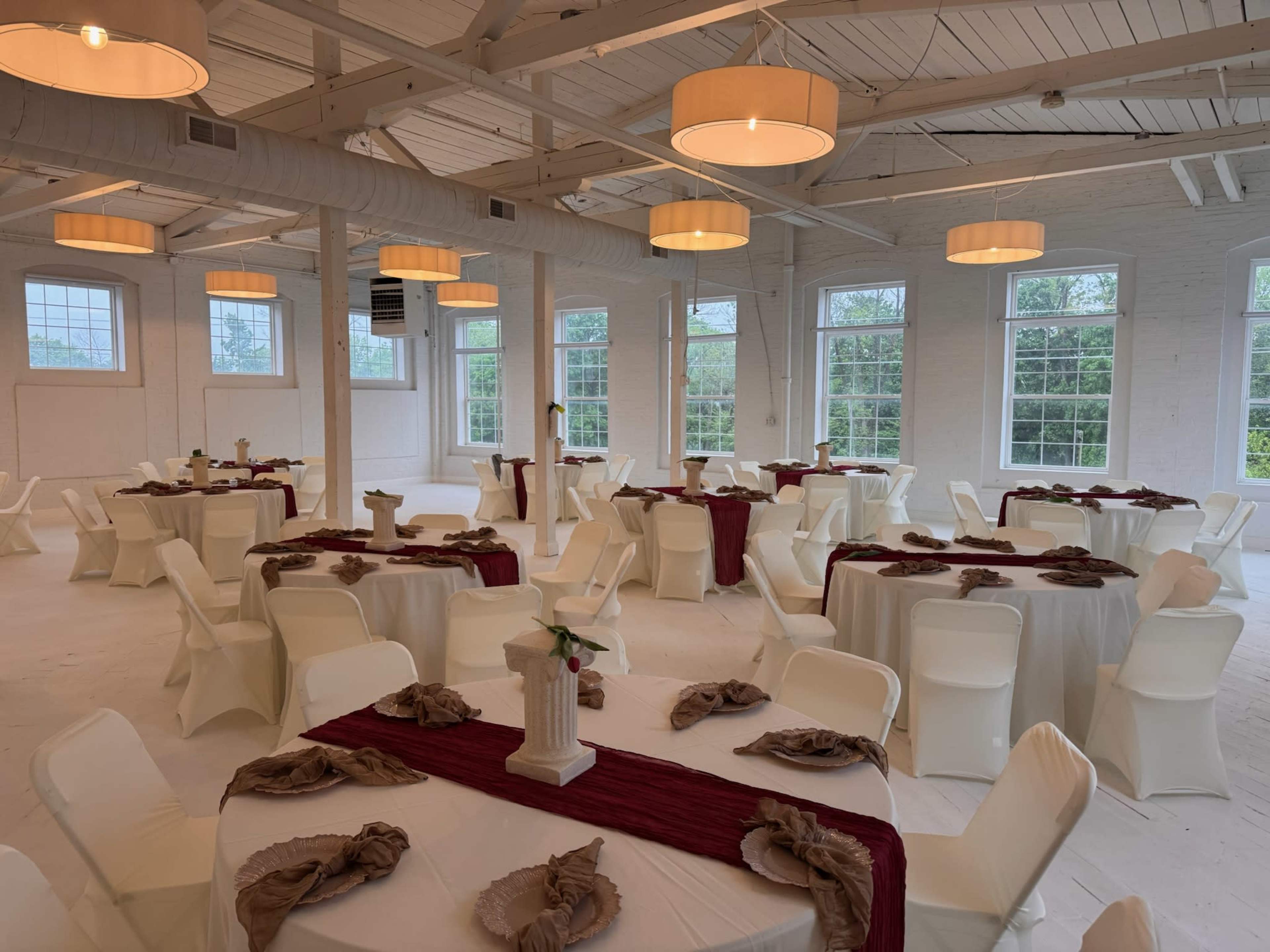 The interior of a spacious event venue features multiple round tables dressed in white tablecloths, red runners, and decorative centerpieces, surrounded by white folding chairs under hanging pendant lights.