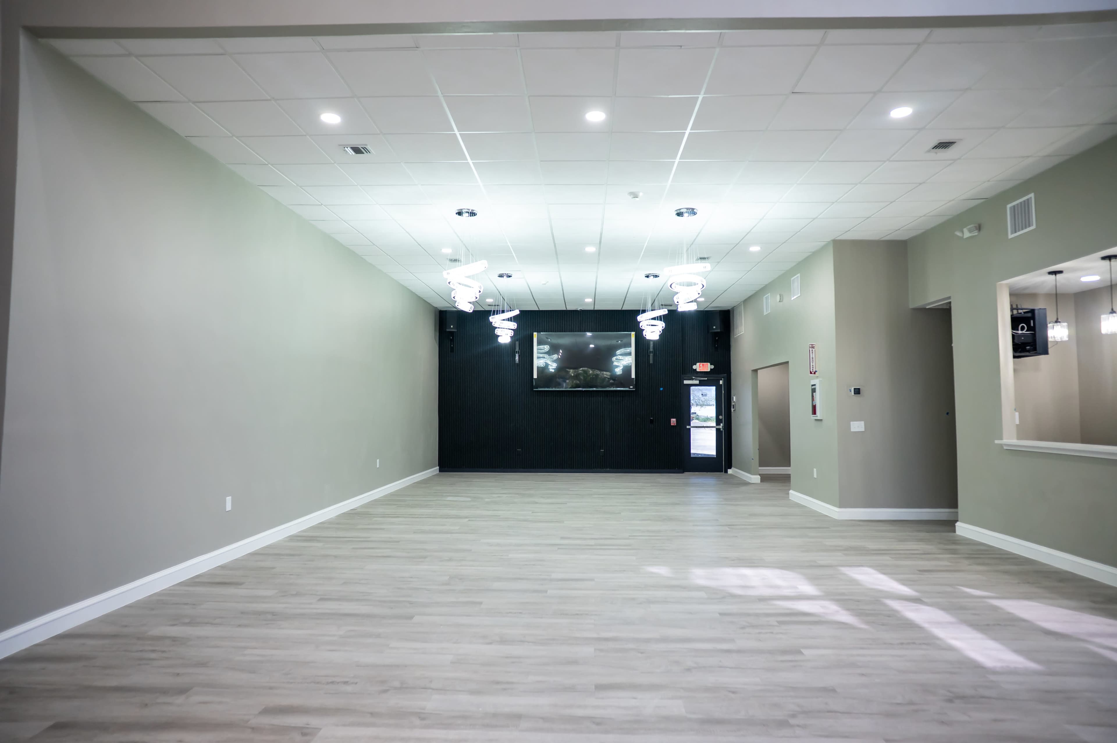The image shows a spacious room with light wood flooring, modern lighting fixtures, and a dark accent wall featuring a mounted screen.
