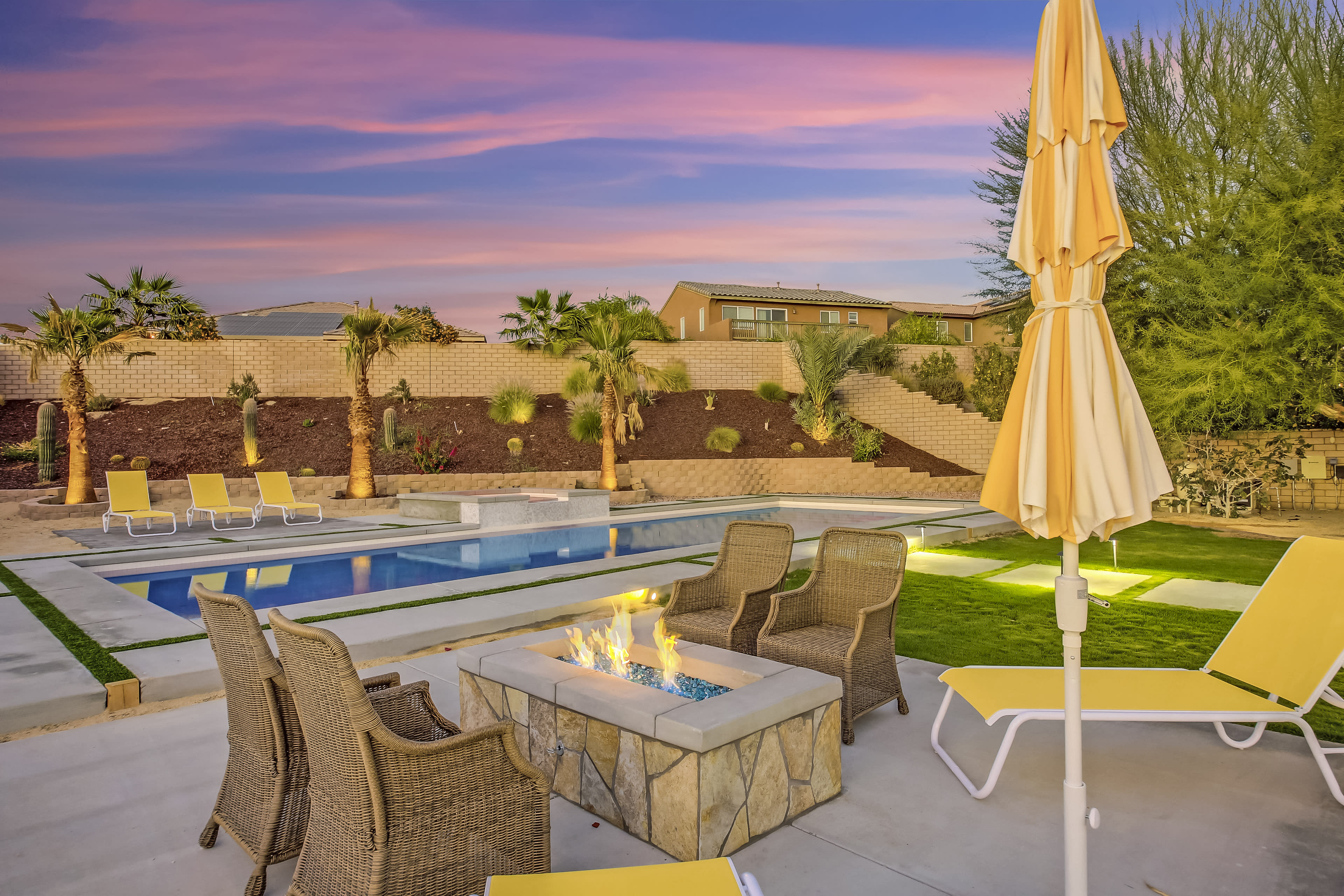 The image depicts a backyard pool area featuring a fire pit surrounded by wicker chairs and yellow lounge chairs under an umbrella, set against a backdrop of palm trees and a colorful sky at sunset.