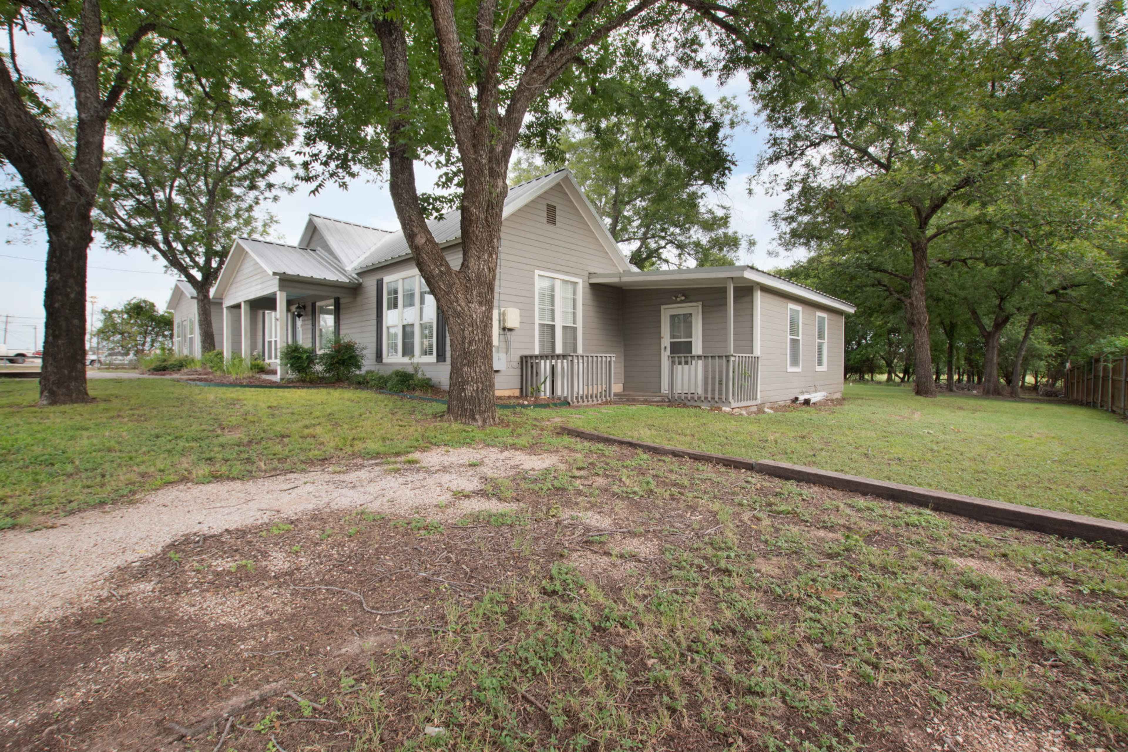 A single-story gray house with a porch is surrounded by trees and a grassy area.