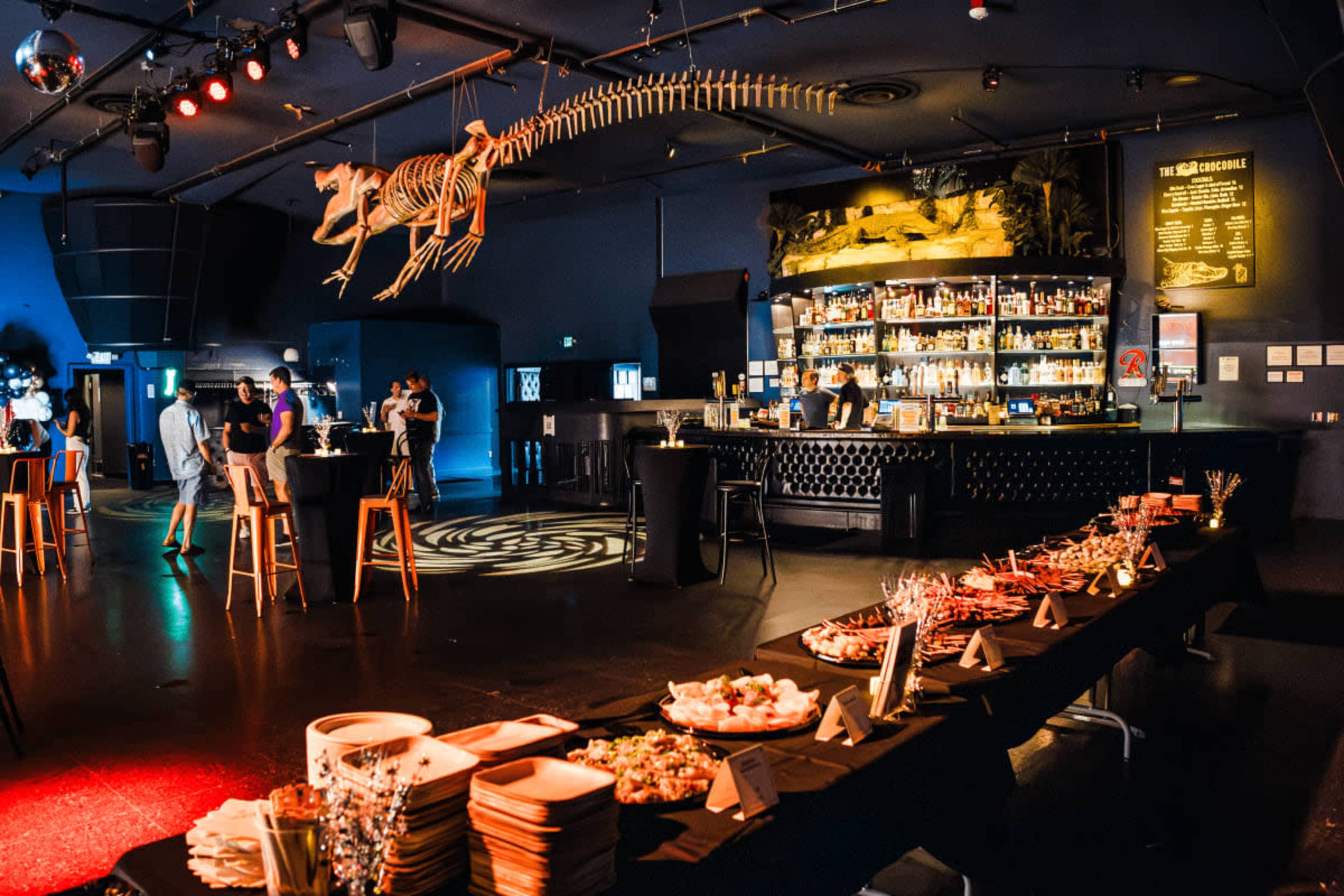 A bar area is set up with a long table displaying various food items, while a skeletal dinosaur hangs from the ceiling in a venue with dim lighting.