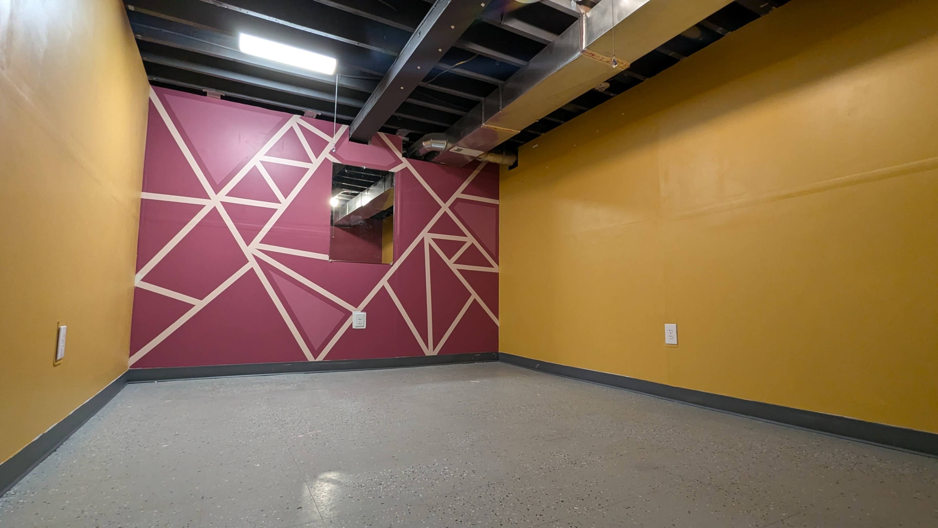 The image shows a small, empty room with yellow and maroon walls featuring geometric designs and a polished gray floor.