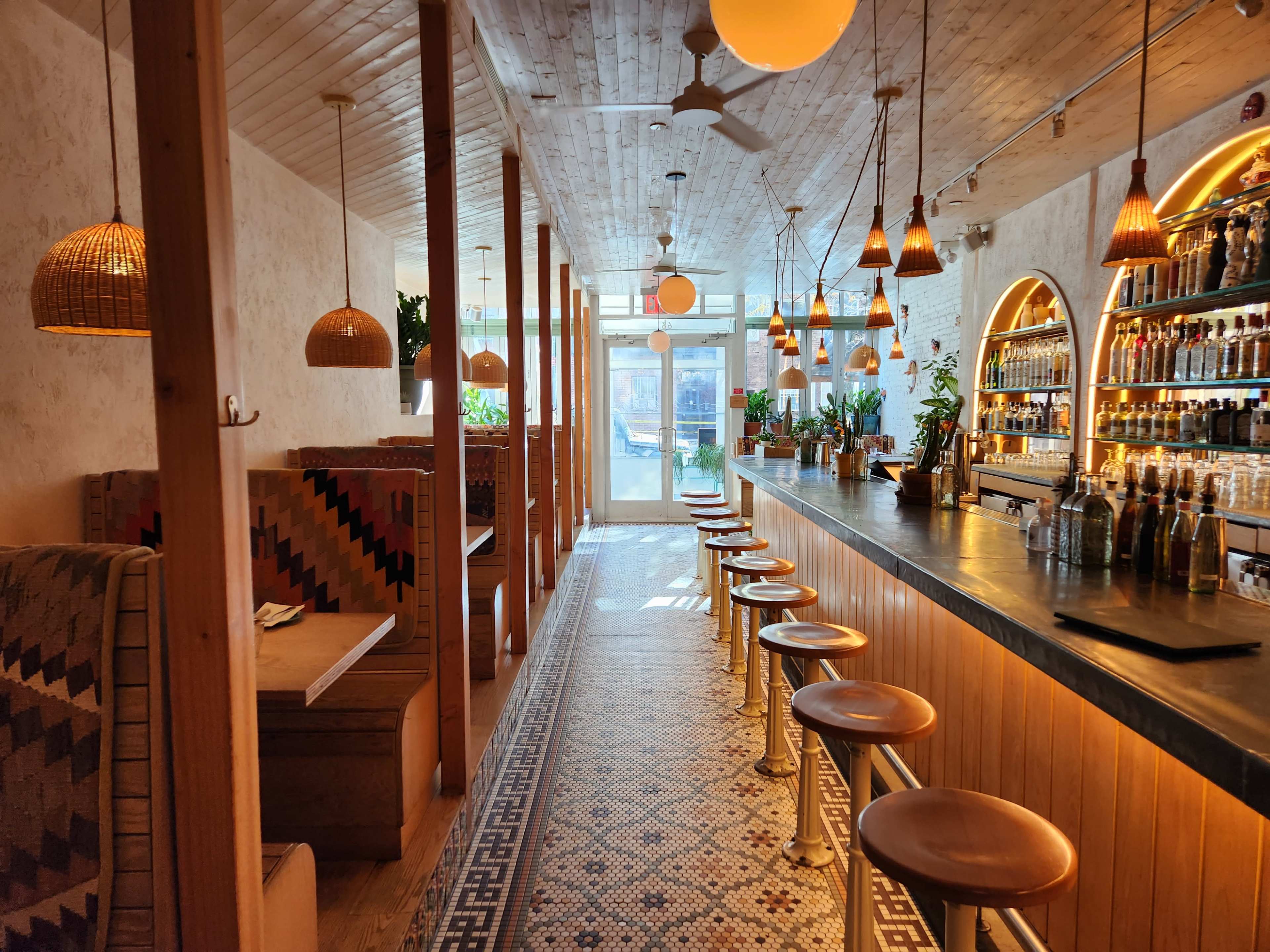 A modern restaurant interior with a long bar, wooden beams, and decorative lighting, featuring seating along one side and a well-stocked bar on the opposite wall.