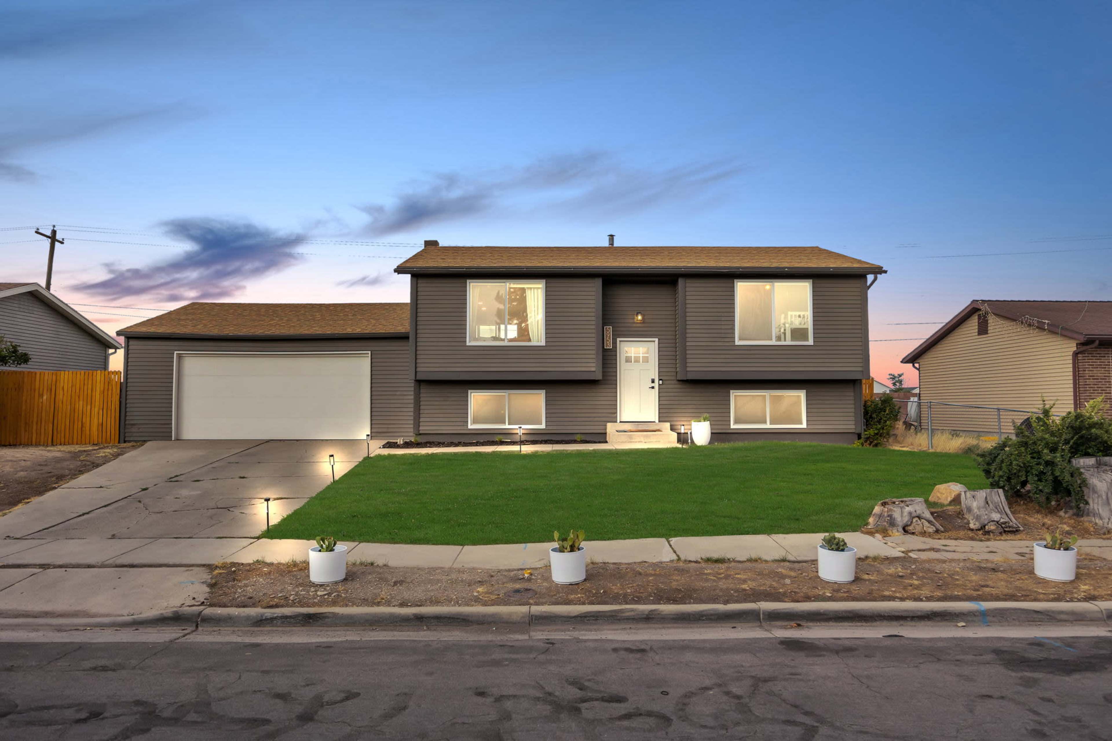 A two-story gray house with a front lawn and a driveway is situated on a suburban street at dusk.