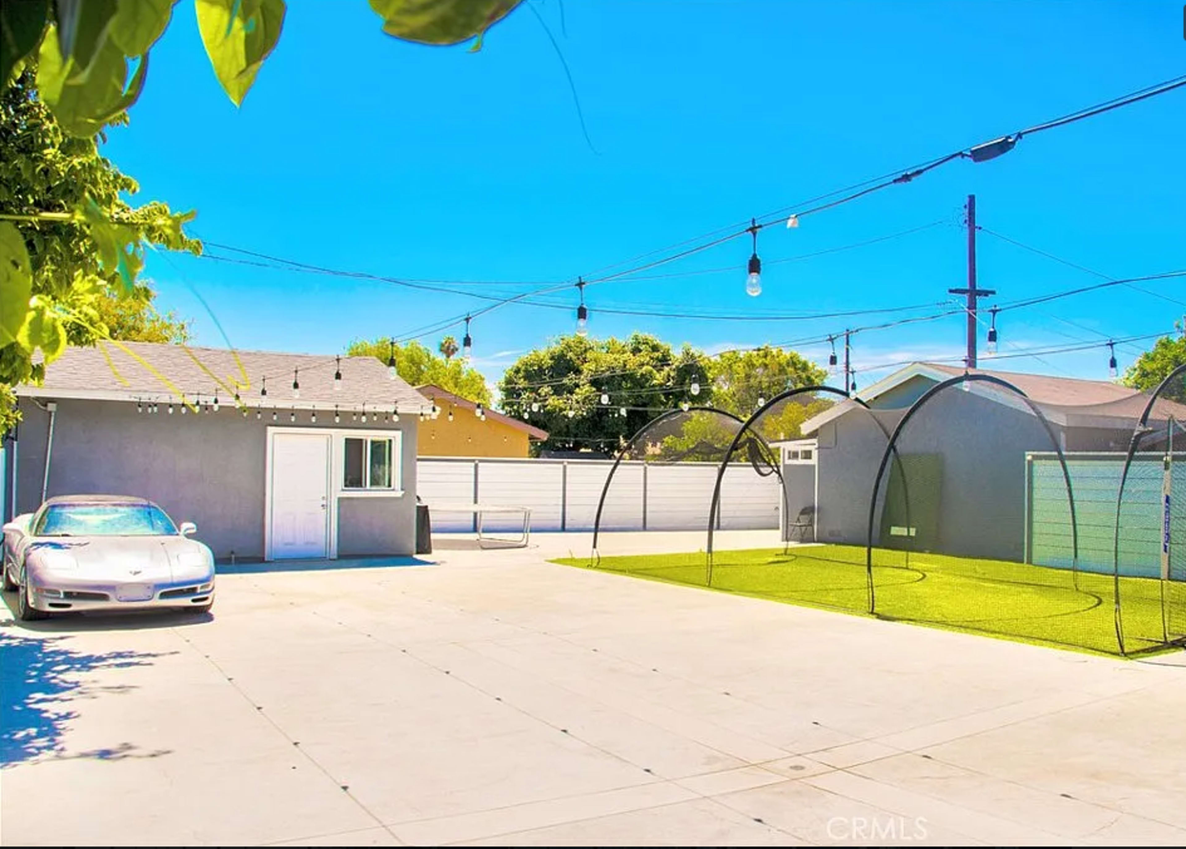 The image shows a spacious outdoor area with a parked silver sports car, a small building, and a fenced grassy section under bright blue skies.