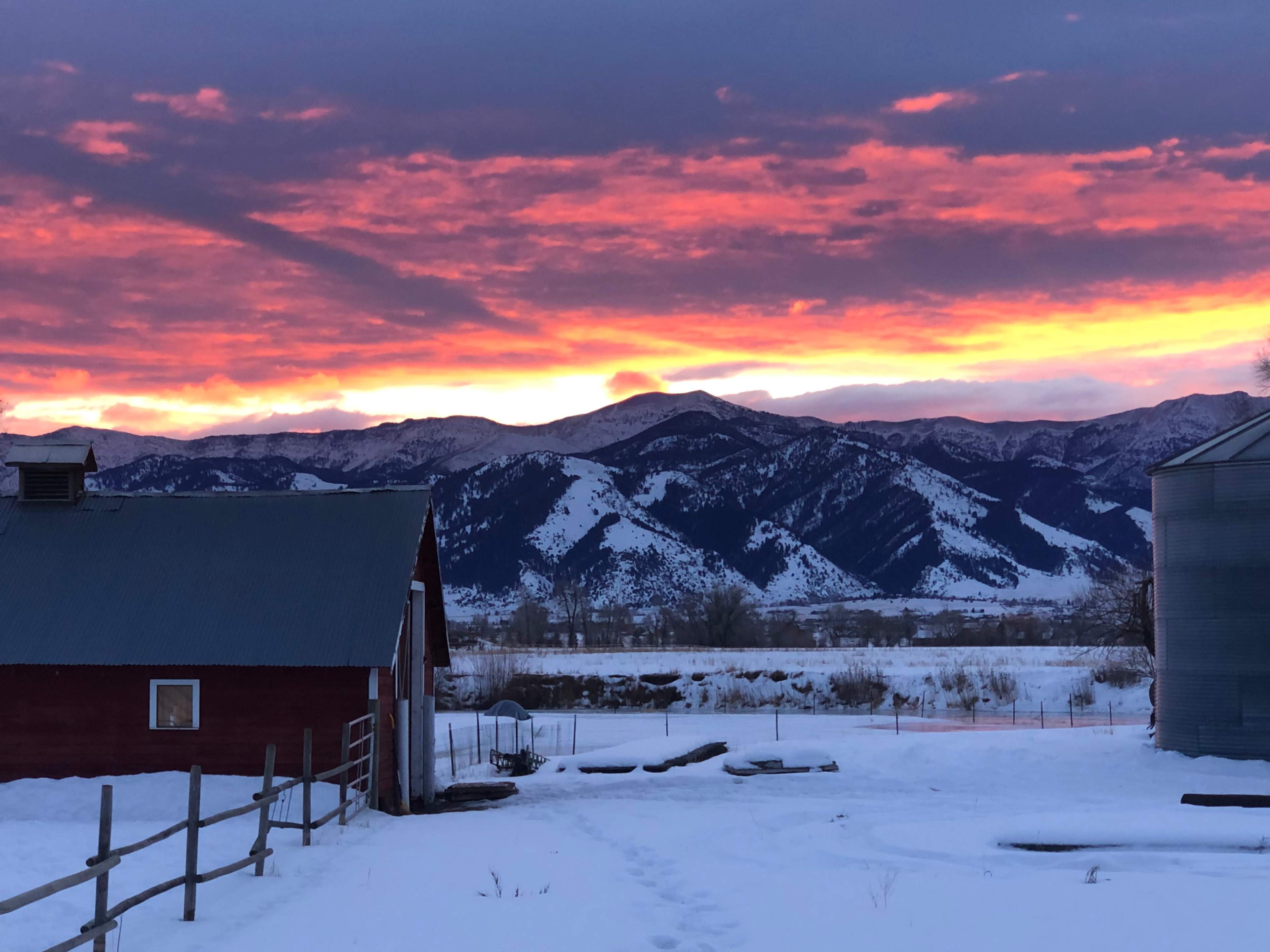 A red barn and a silos are set against snow-covered ground and mountains under a colorful sunset sky.