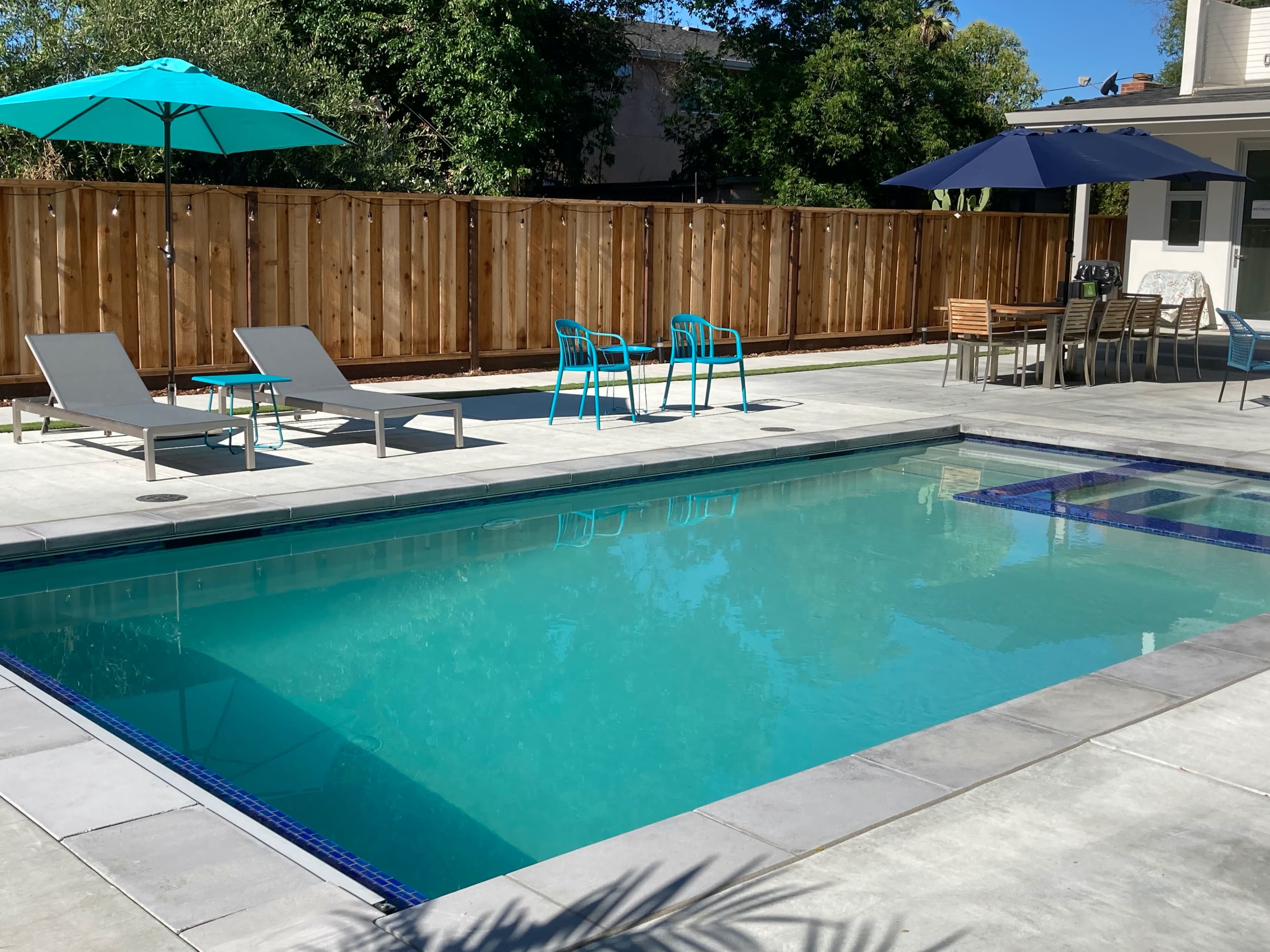 A clear swimming pool surrounded by lounge chairs, outdoor tables, and umbrellas in a fenced backyard.