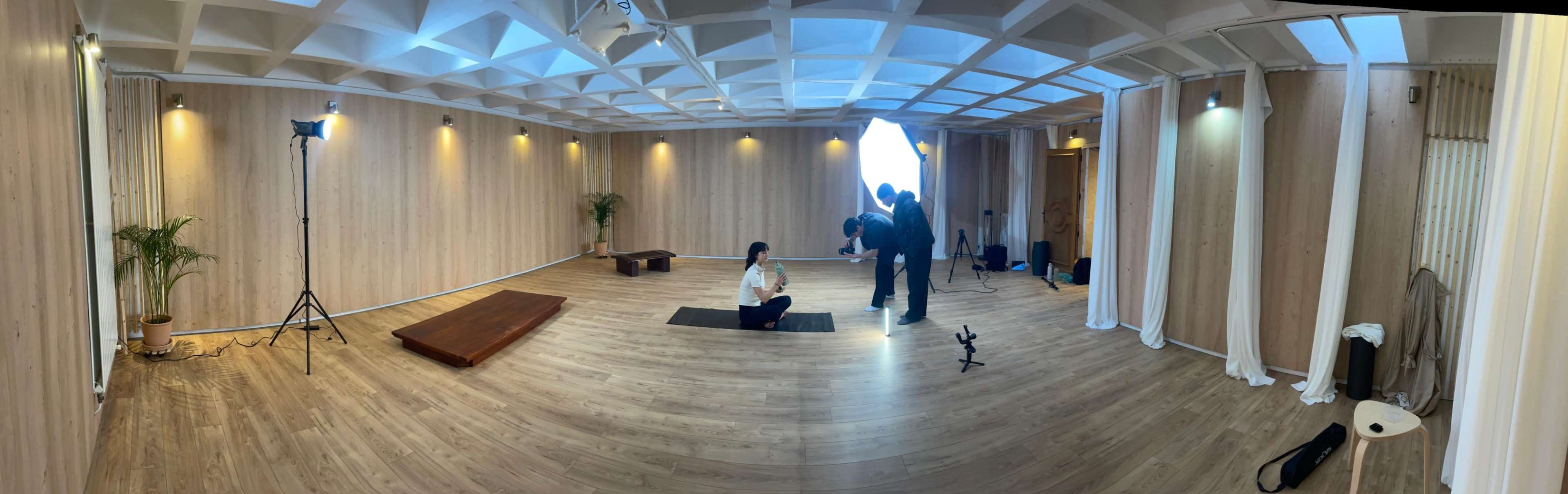 The image shows a spacious, well-lit interior studio with wooden flooring, featuring a person seated on a yoga mat, while two individuals prepare equipment nearby.