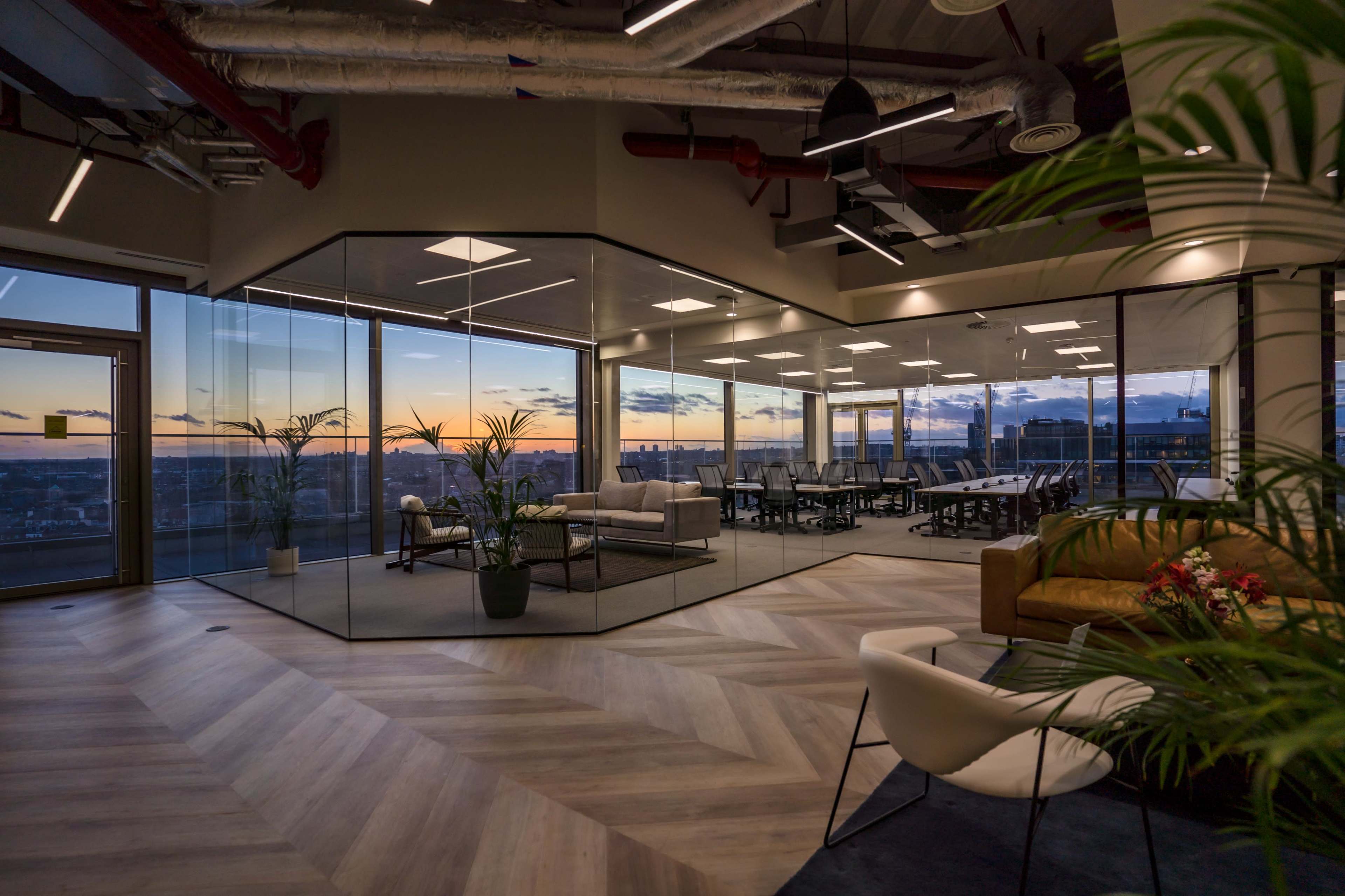 A modern office space featuring a glass-walled meeting area and a view of a sunset over the city.