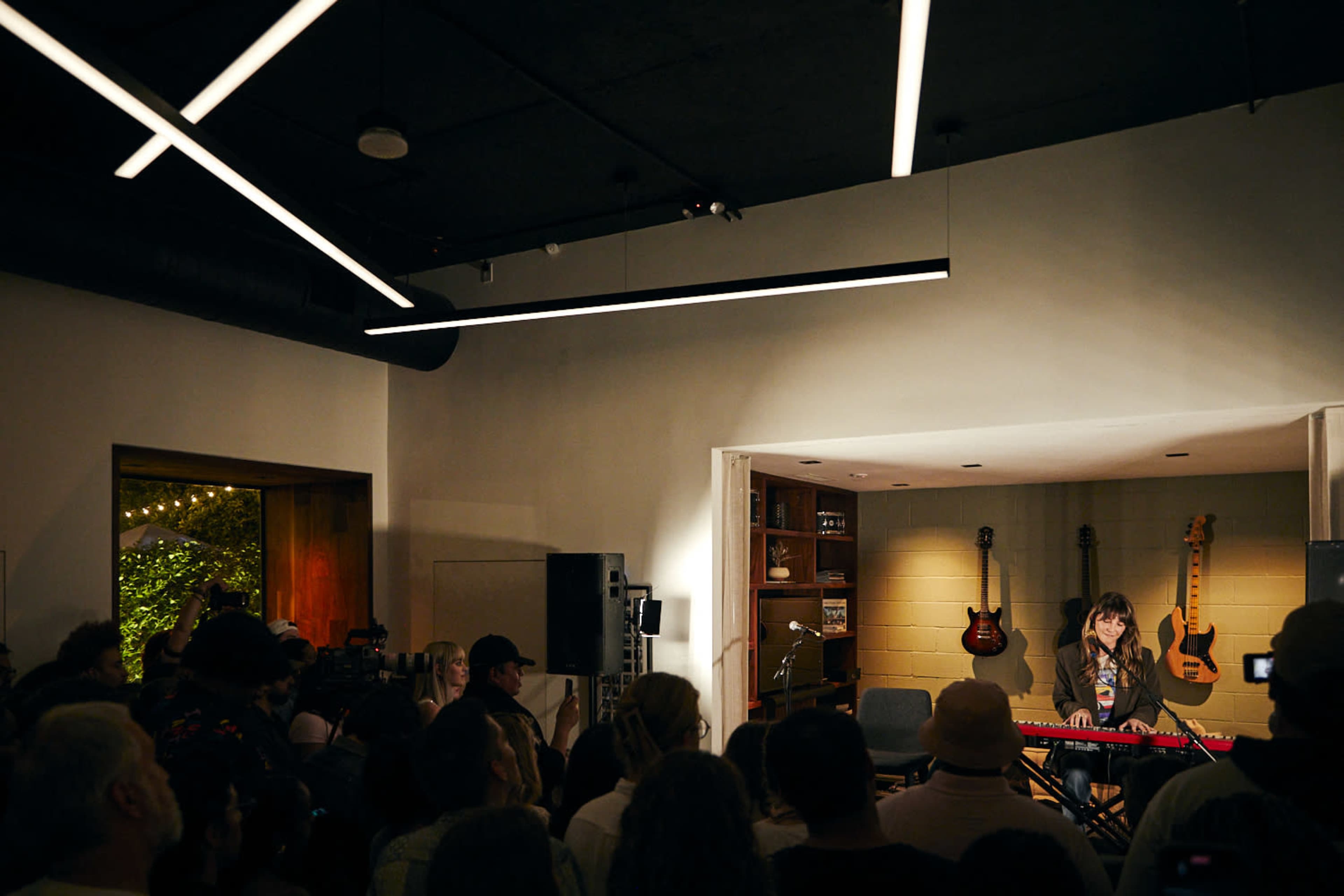 A musician plays a keyboard in a small venue filled with an audience, while guitars adorn the walls behind her.