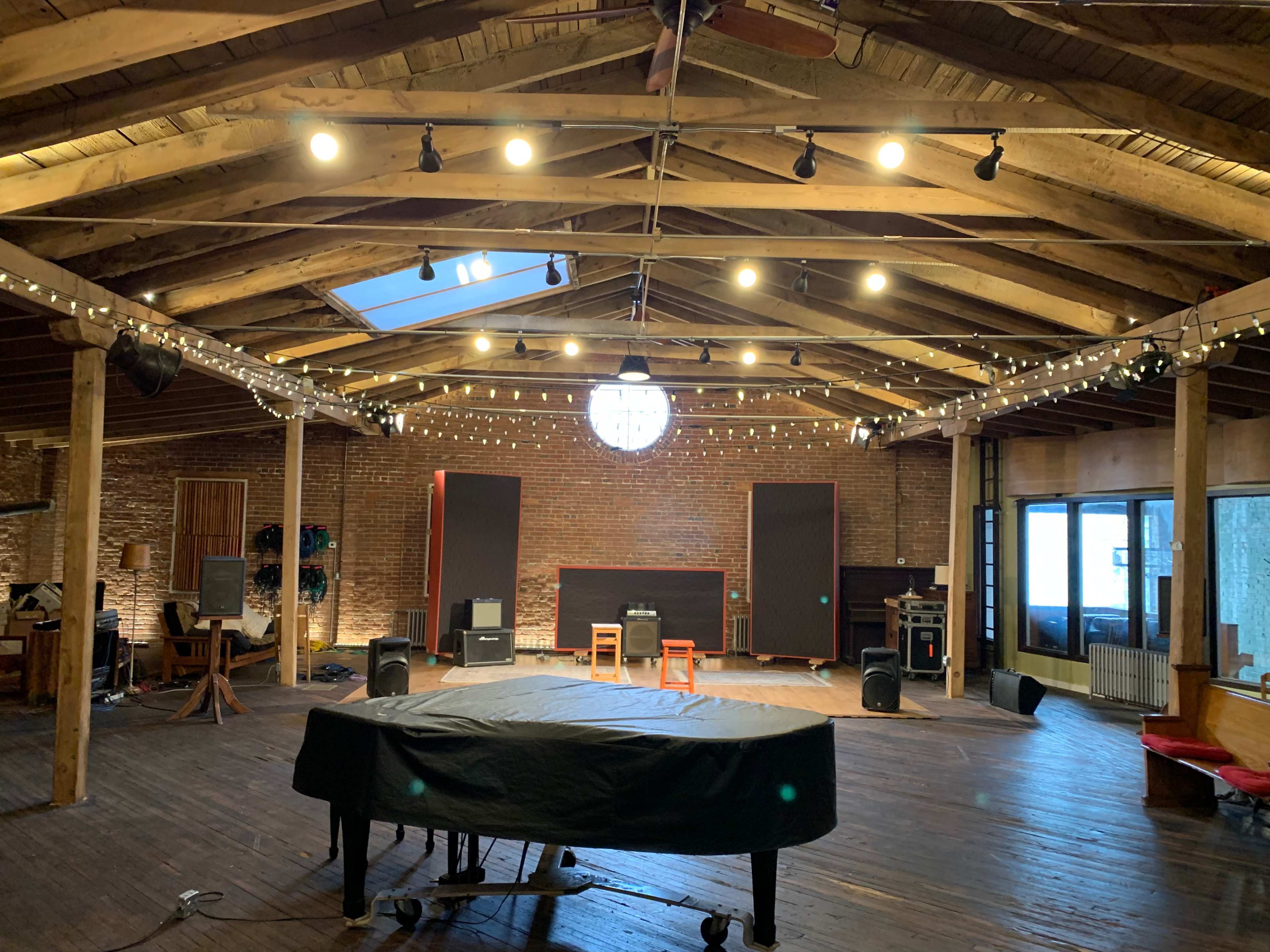 An empty room with high wooden ceilings, a grand piano covered with a cloth, and sound equipment arranged on the wooden floor.
