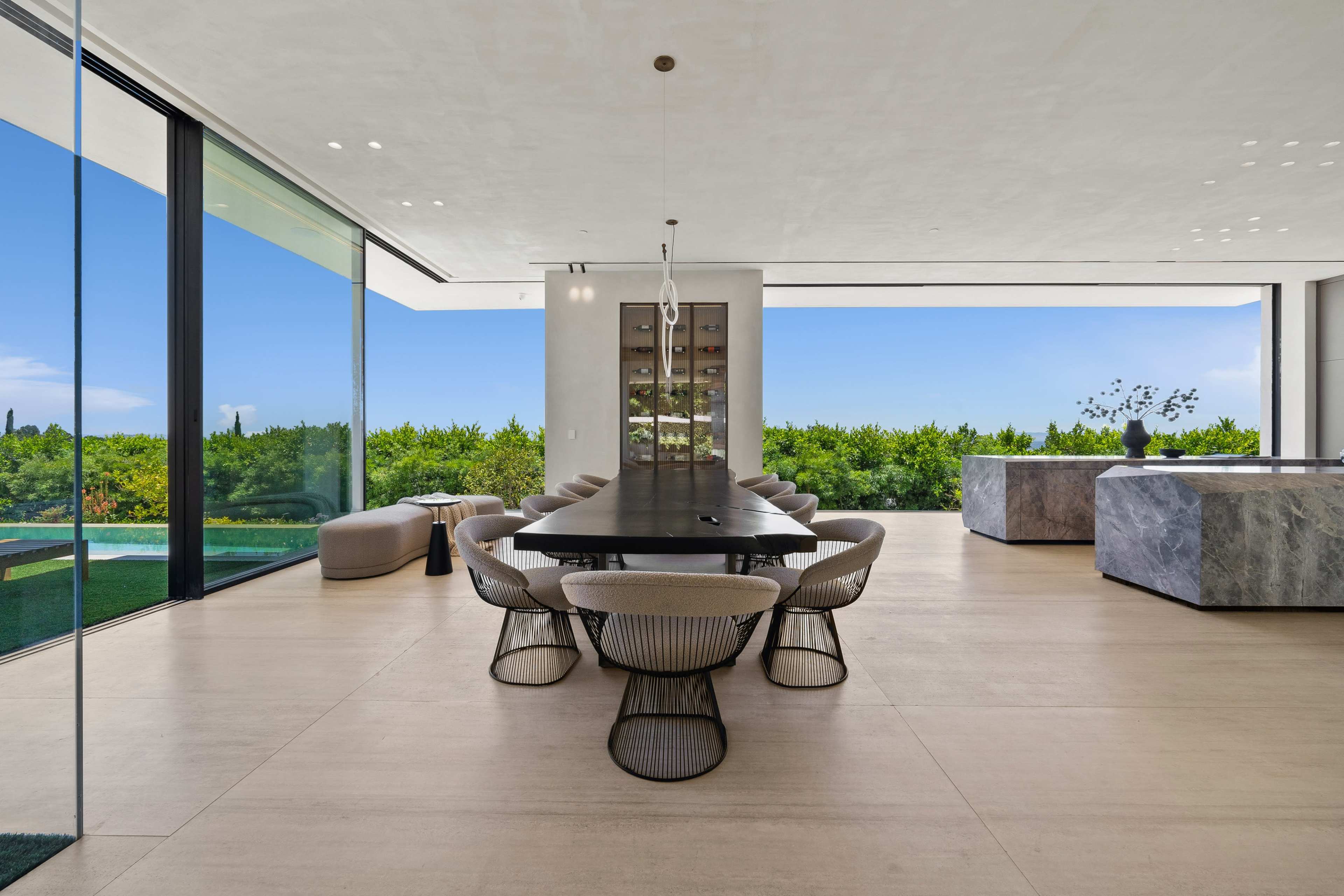 The image shows a modern dining area with a large table surrounded by chairs, featuring floor-to-ceiling windows that overlook a green landscape.