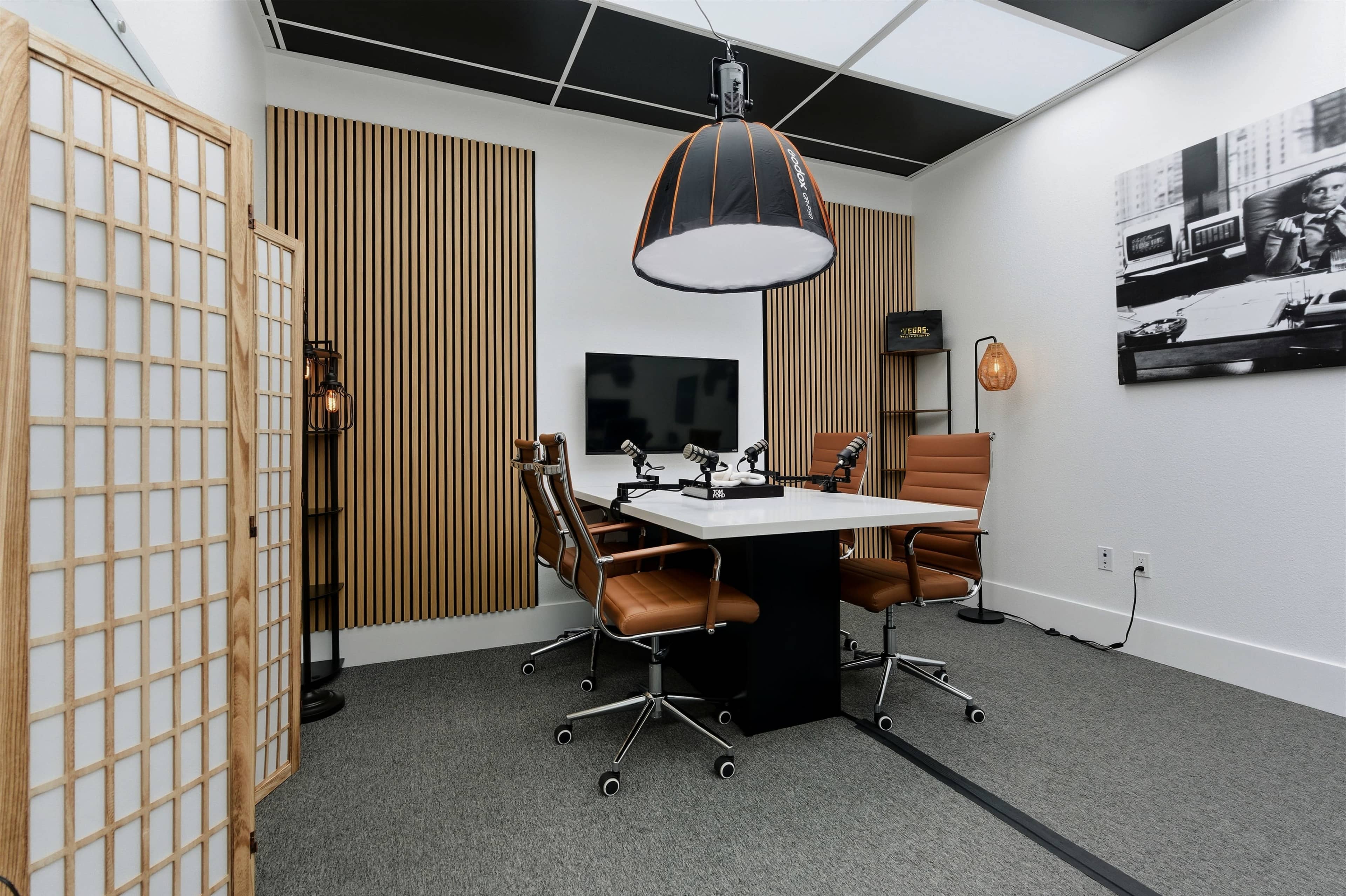The image shows a modern conference room furnished with an elegant table, leather chairs, and wooden accent walls, featuring a large screen and multiple light fixtures.