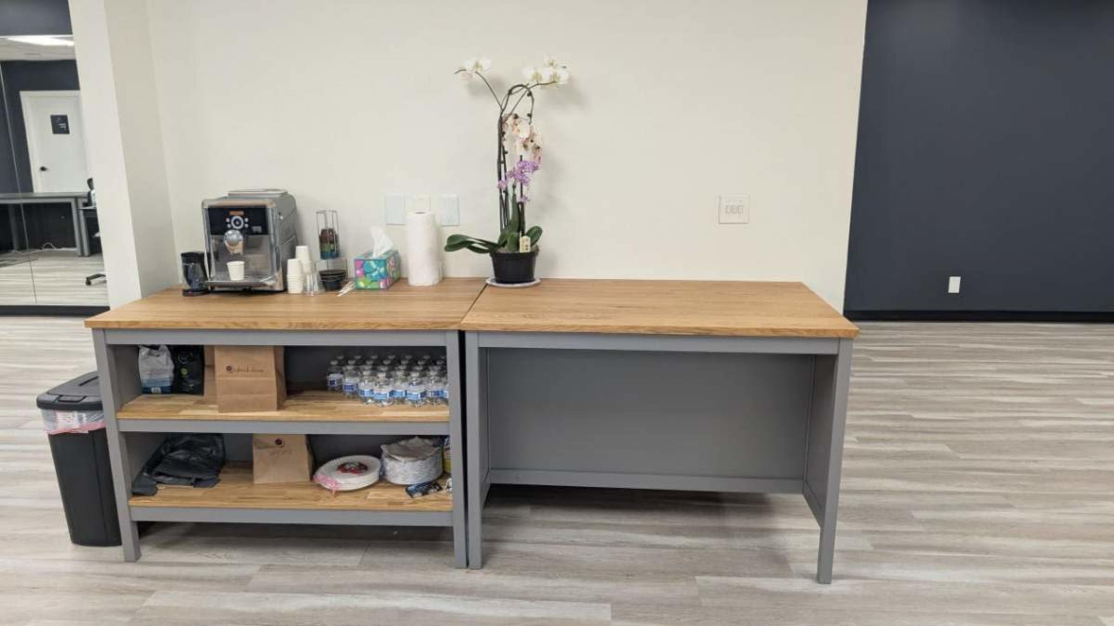 A wooden table with a grey base holds a coffee maker, cups, and a potted orchid, while the lower shelf contains bottled water and paper plates.