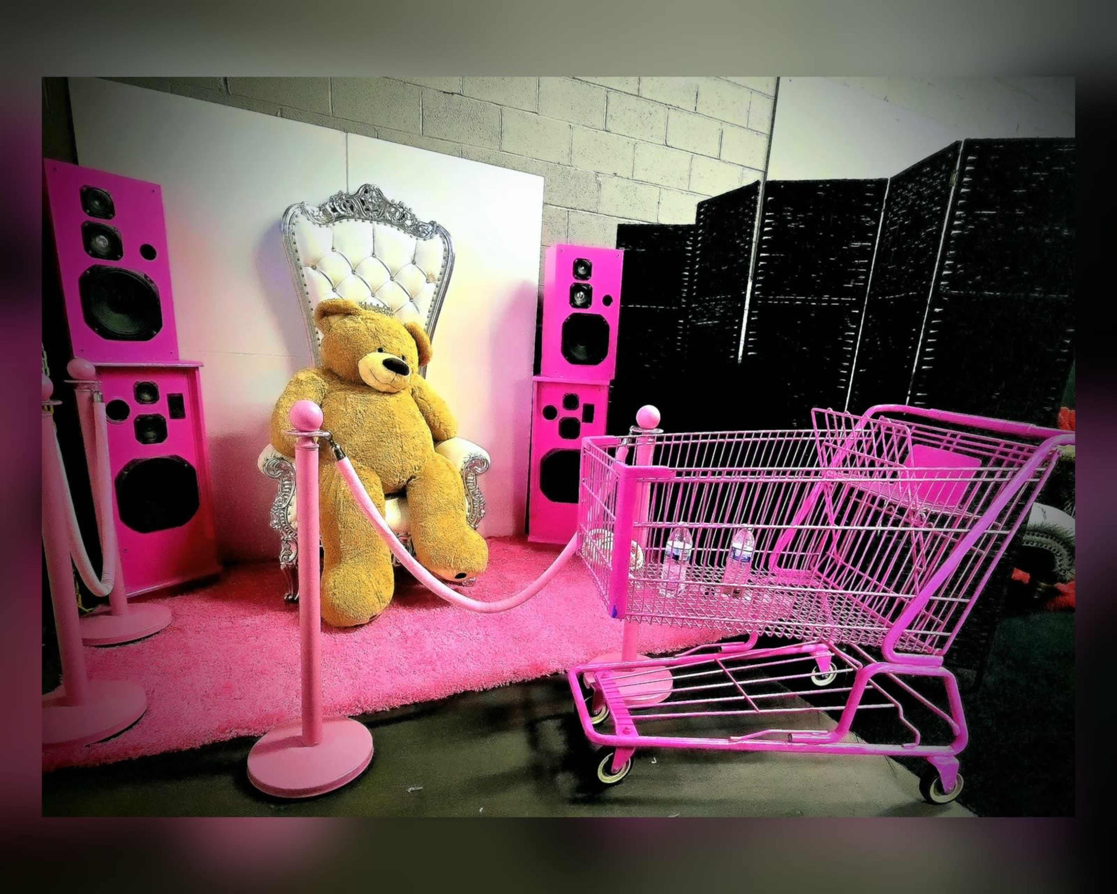 The scene features a large teddy bear sitting on a ornate throne, surrounded by pink speakers, with a pink shopping cart nearby.