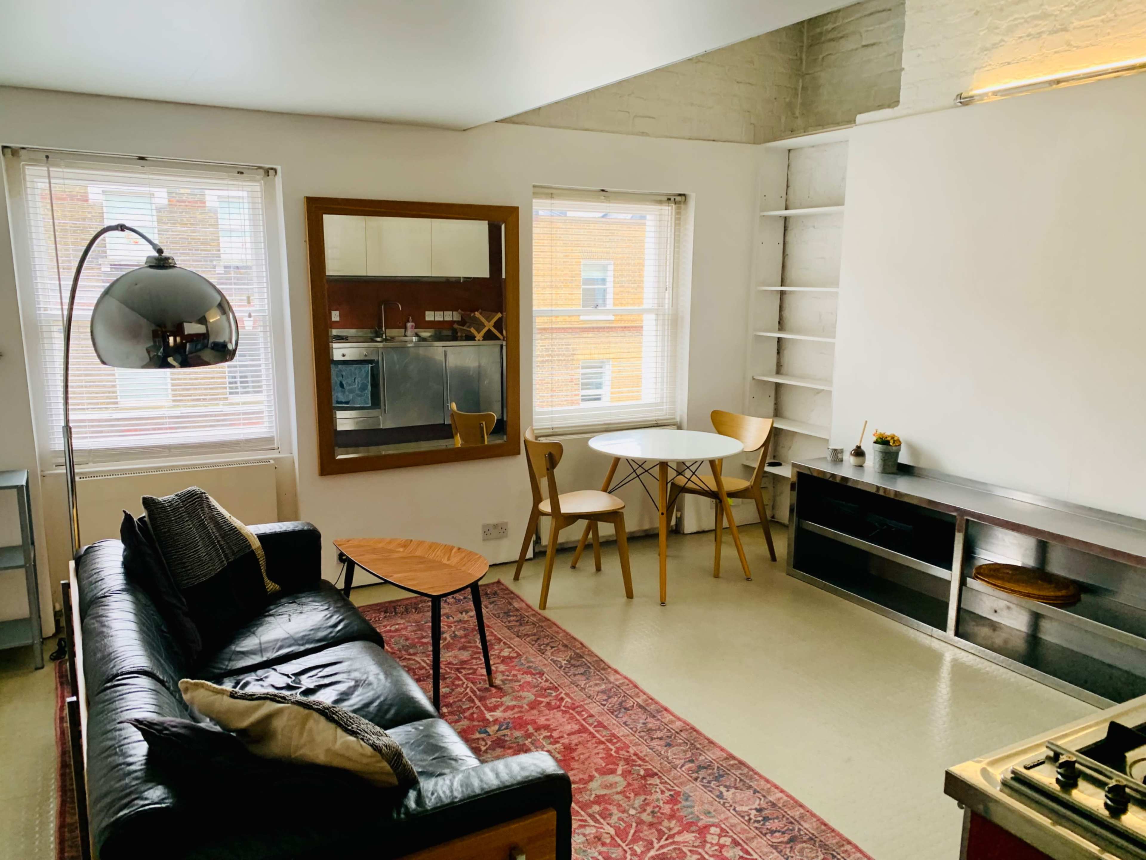 The image shows a modern living area featuring a black leather sofa, a round dining table with chairs, and shelves along the wall in a well-lit room.
